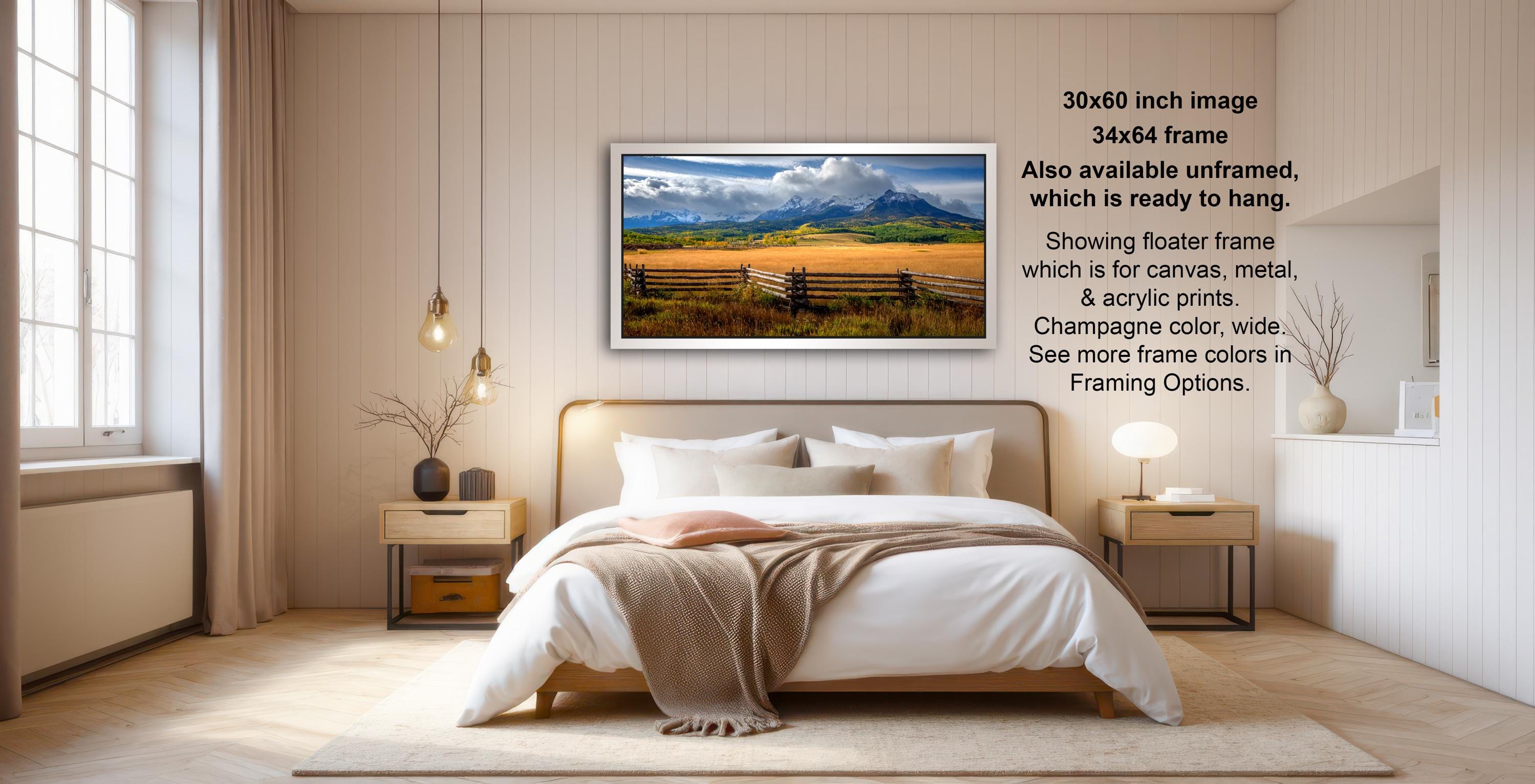 The image is a bedroom with a large bed, wooden furniture, and a framed landscape photograph hanging on the wall. The photograph depicts a scenic countryside landscape with a mountain range in the background. The bedroom has a minimalist and modern design, with a neutral color palette and clean lines.
