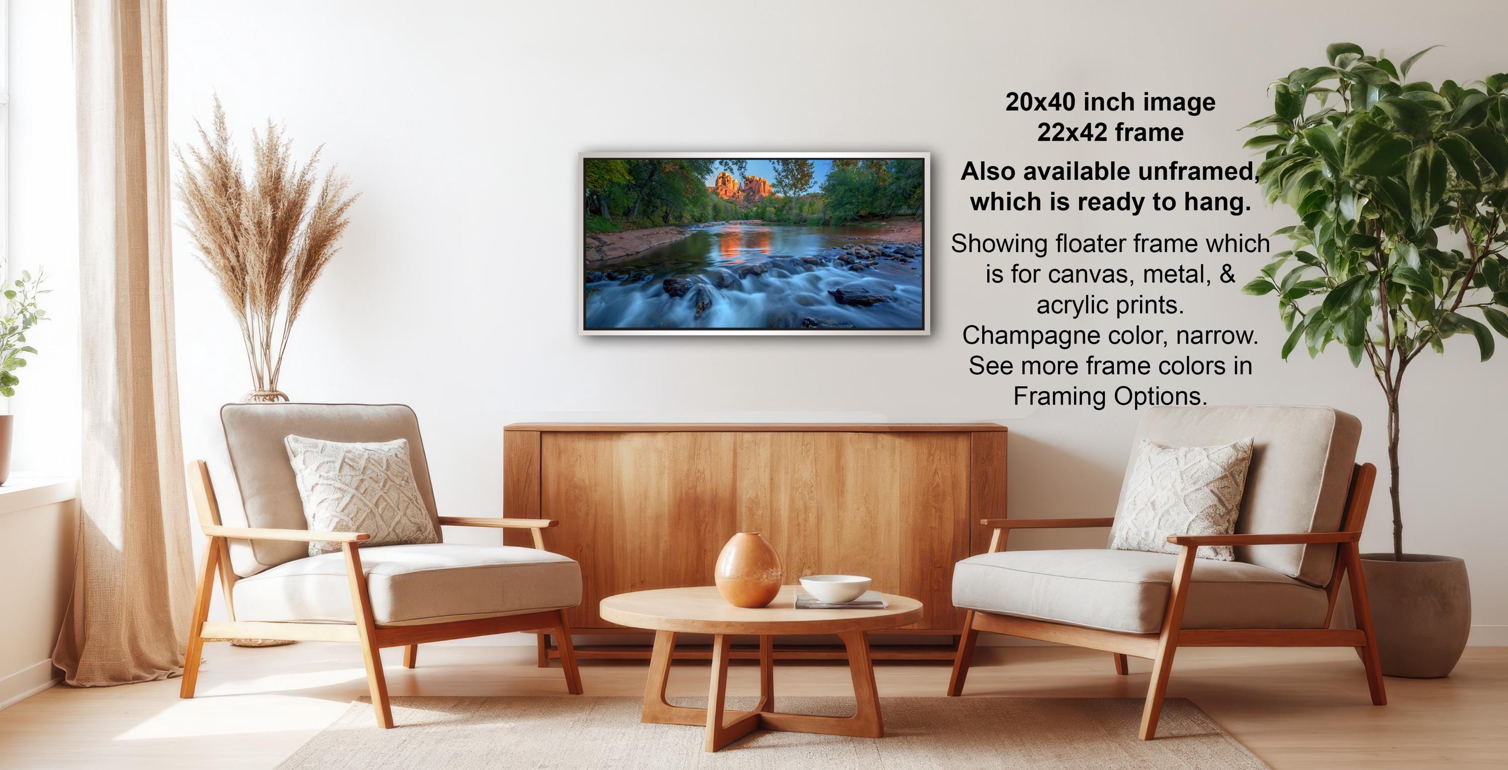 a living room with a wooden coffee table, two white armchairs, and a potted plant. A framed landscape photograph is hanging on the wall, and there is text providing information about the frame options.