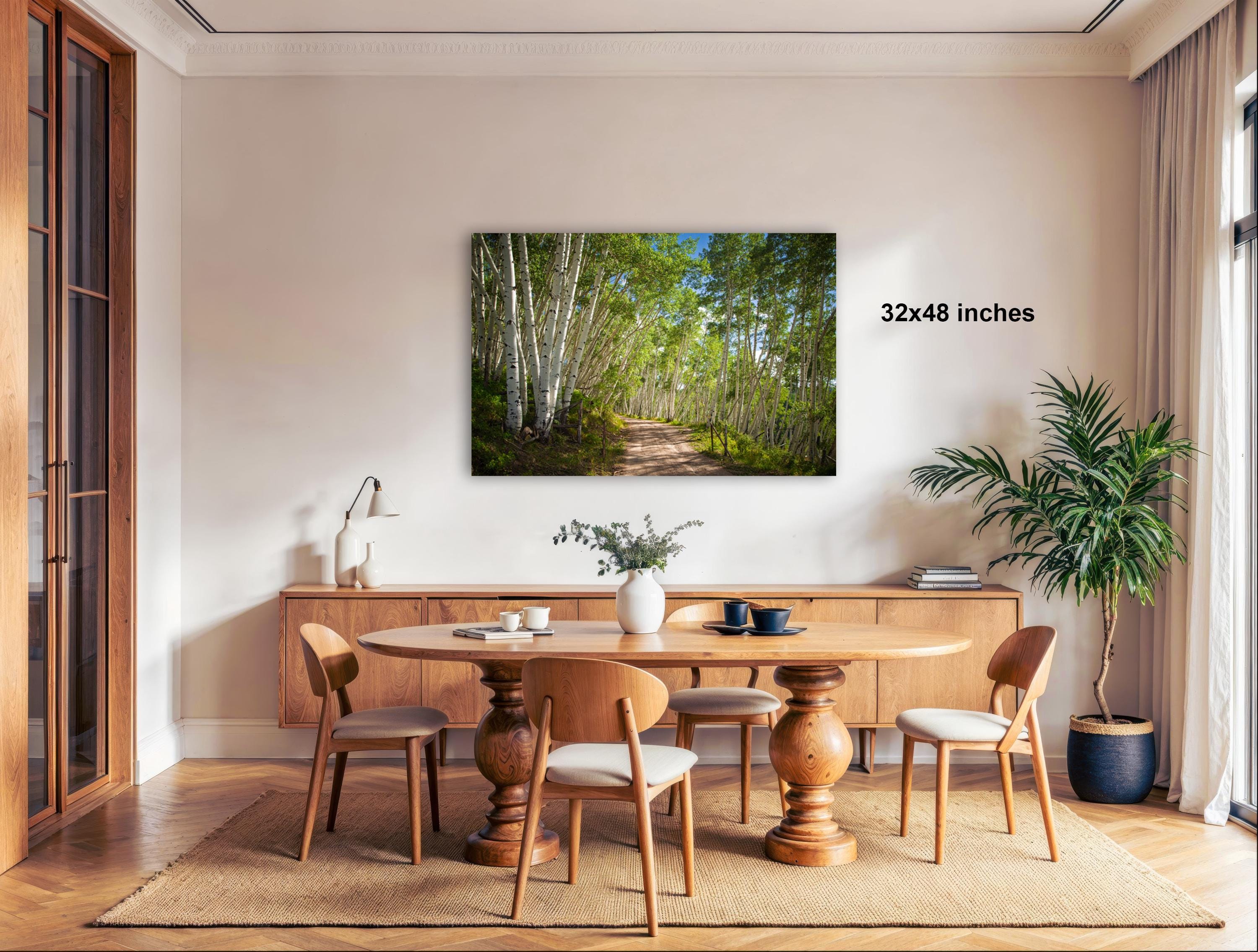 a serene dining room with a large wooden table surrounded by matching chairs. A painting of a forest path is displayed on the wall above the table, adding a natural element to the space. The room is well-lit, with natural light coming in from a window on the right side. The overall atmosphere is calm and inviting, with a touch of greenery from the potted plant and the vase of flowers on the table.