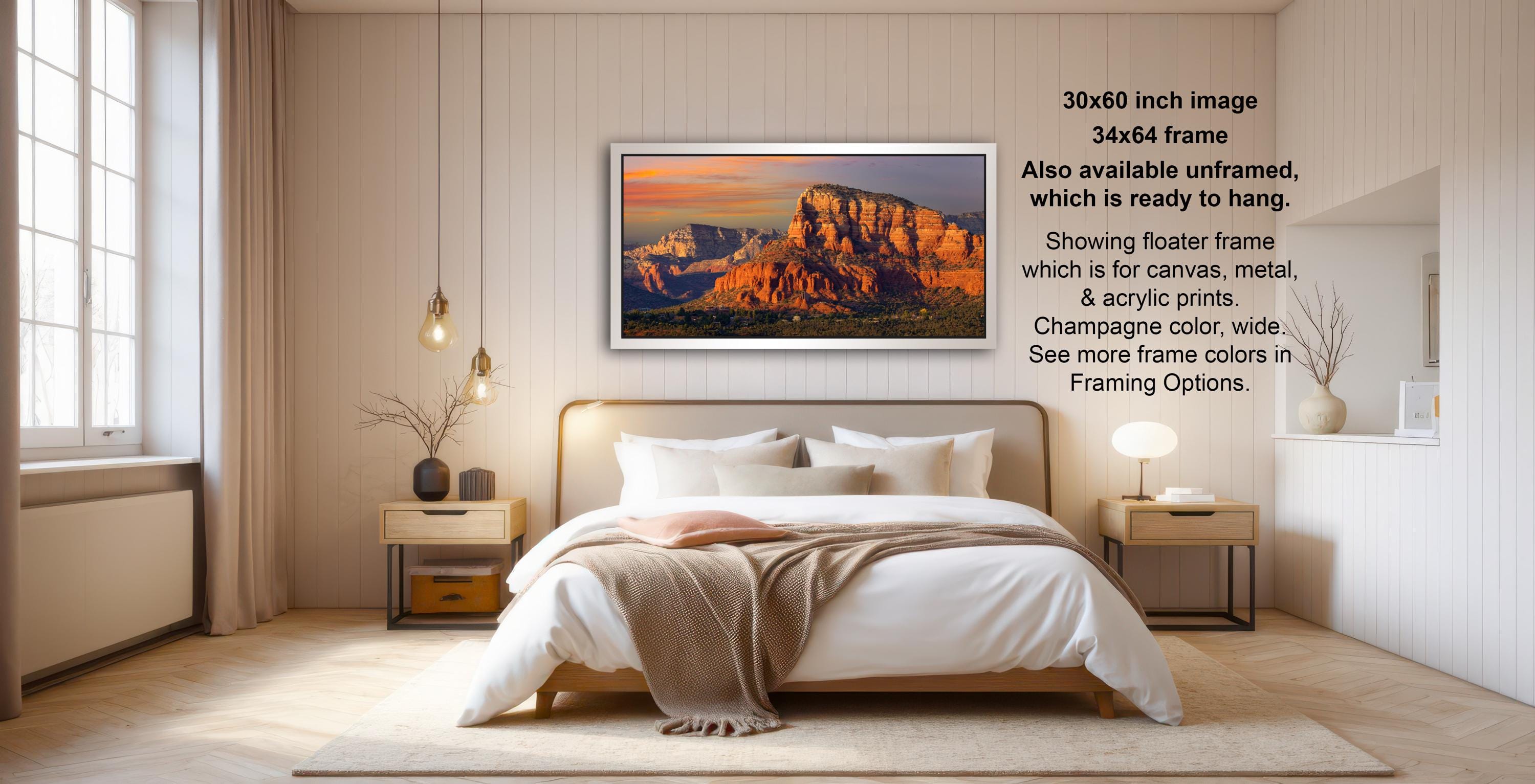 The image is a bedroom with a large bed, a nightstand, and a painting of a mountain landscape hanging on the wall. The painting is framed in a wooden frame and is ready to hang. The room has a minimalist and modern design, with a white color scheme and a neutral color palette.