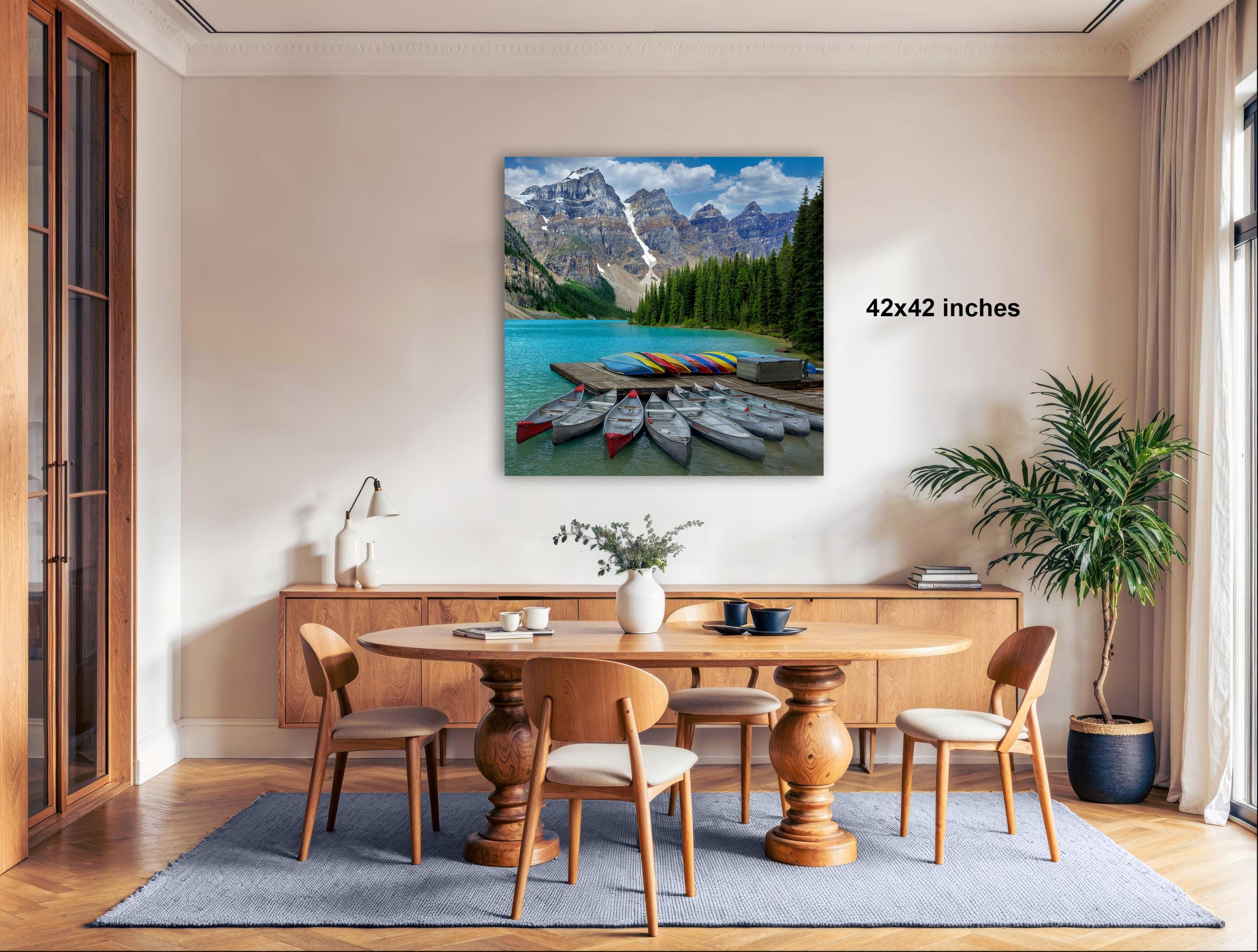 a dining room with a large wooden table surrounded by chairs, a potted plant, and a painting of a mountain lake with boats on the shore.