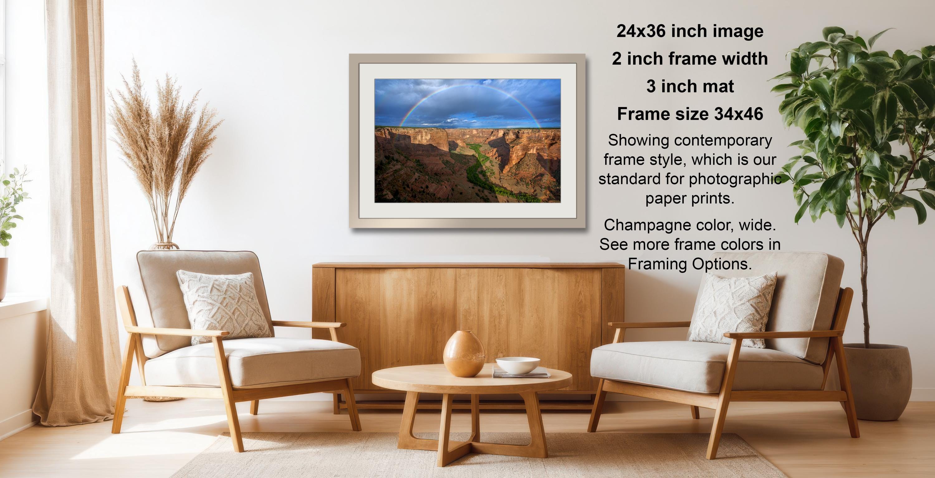 a living room with a wooden coffee table, two white armchairs, and a framed photograph of a desert landscape with a rainbow. The photograph is framed in a 2x3 grid and has a size of 34x46 inches. The frame style is described as contemporary, and the photograph is showing the standard for photographic paper prints. The color of the frame is champagne, and there are options to see more frame colors in Framing Options.