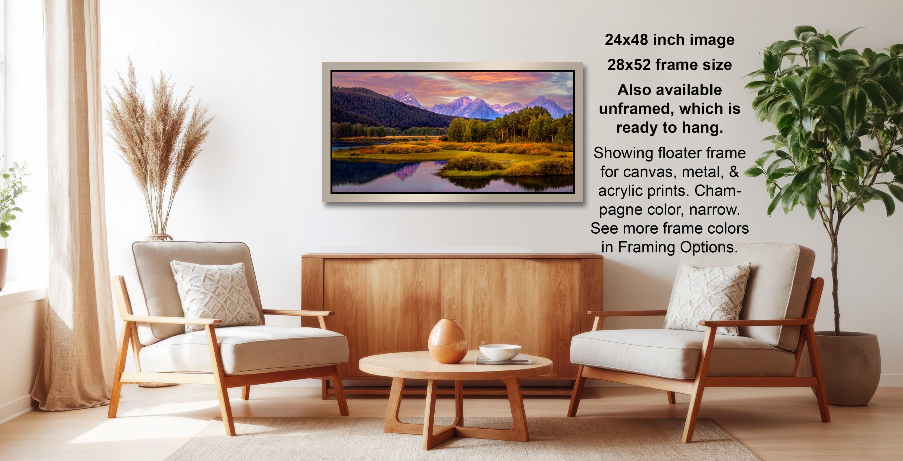 a living room with a large framed landscape painting hanging on the wall. The painting depicts a serene mountain landscape with a lake and trees. The room is furnished with two chairs and a coffee table, creating a cozy and inviting atmosphere.