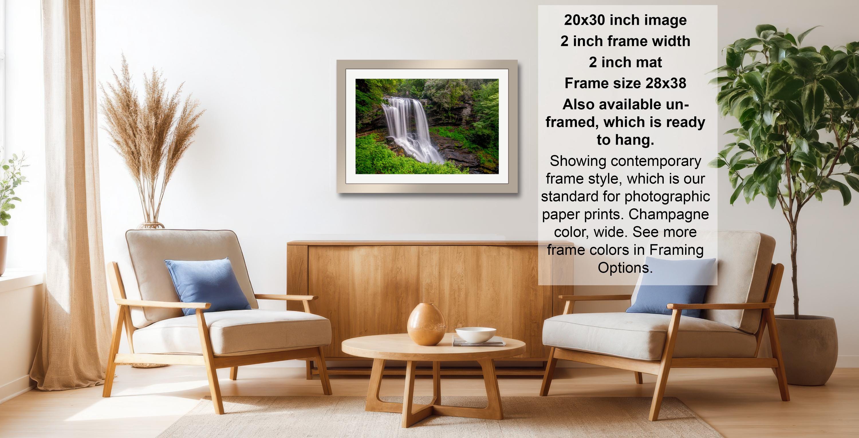 a living room with a framed photograph of a waterfall hanging on the wall. The room has two armchairs, a coffee table, and a potted plant. The photograph is framed in a modern style, and the frame size is 28.38 inches wide. The text indicates that the frame is available in both framed and unframed versions, and it is ready to hang.