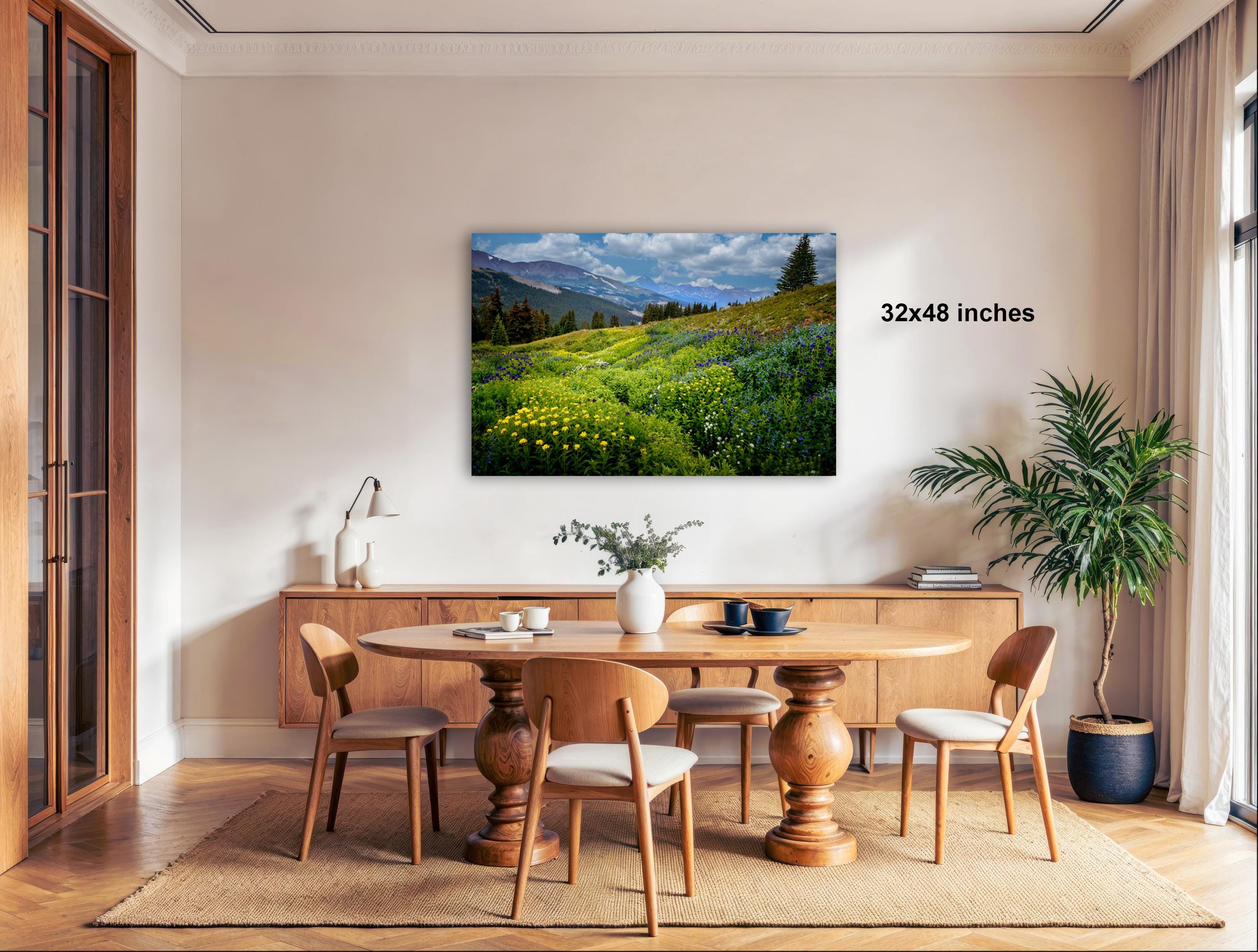 a serene dining room with a large wooden table surrounded by matching chairs. A painting of a lush green field with yellow flowers hangs on the wall above the table. The room is decorated with potted plants and other natural elements, creating a peaceful and inviting atmosphere.