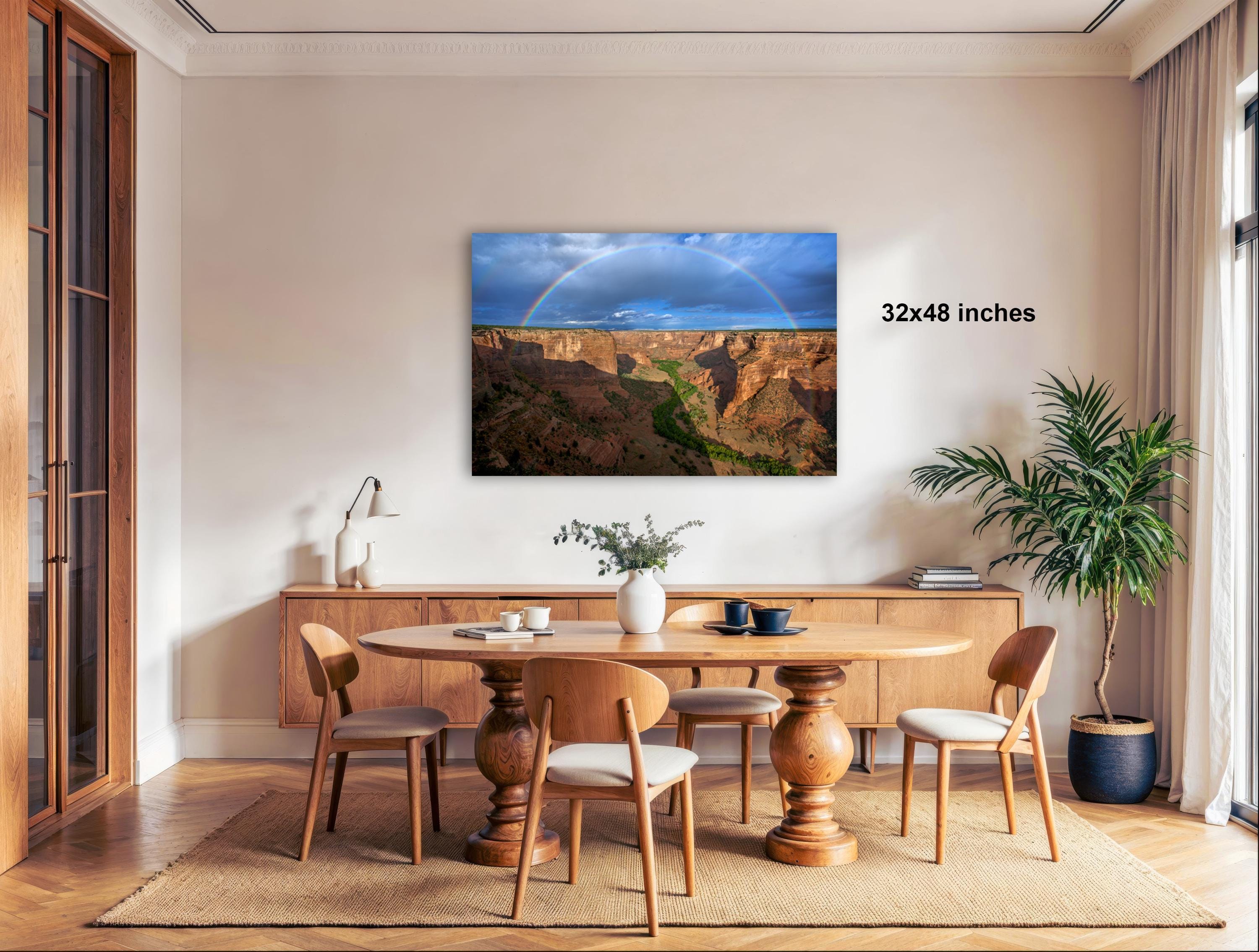a large wooden dining table surrounded by wooden chairs, with a vase of flowers and a potted plant on the table. Above the table, there is a large framed picture of a mountain landscape with a rainbow in the sky. The wall behind the table has a text that reads "32x48 inches".
