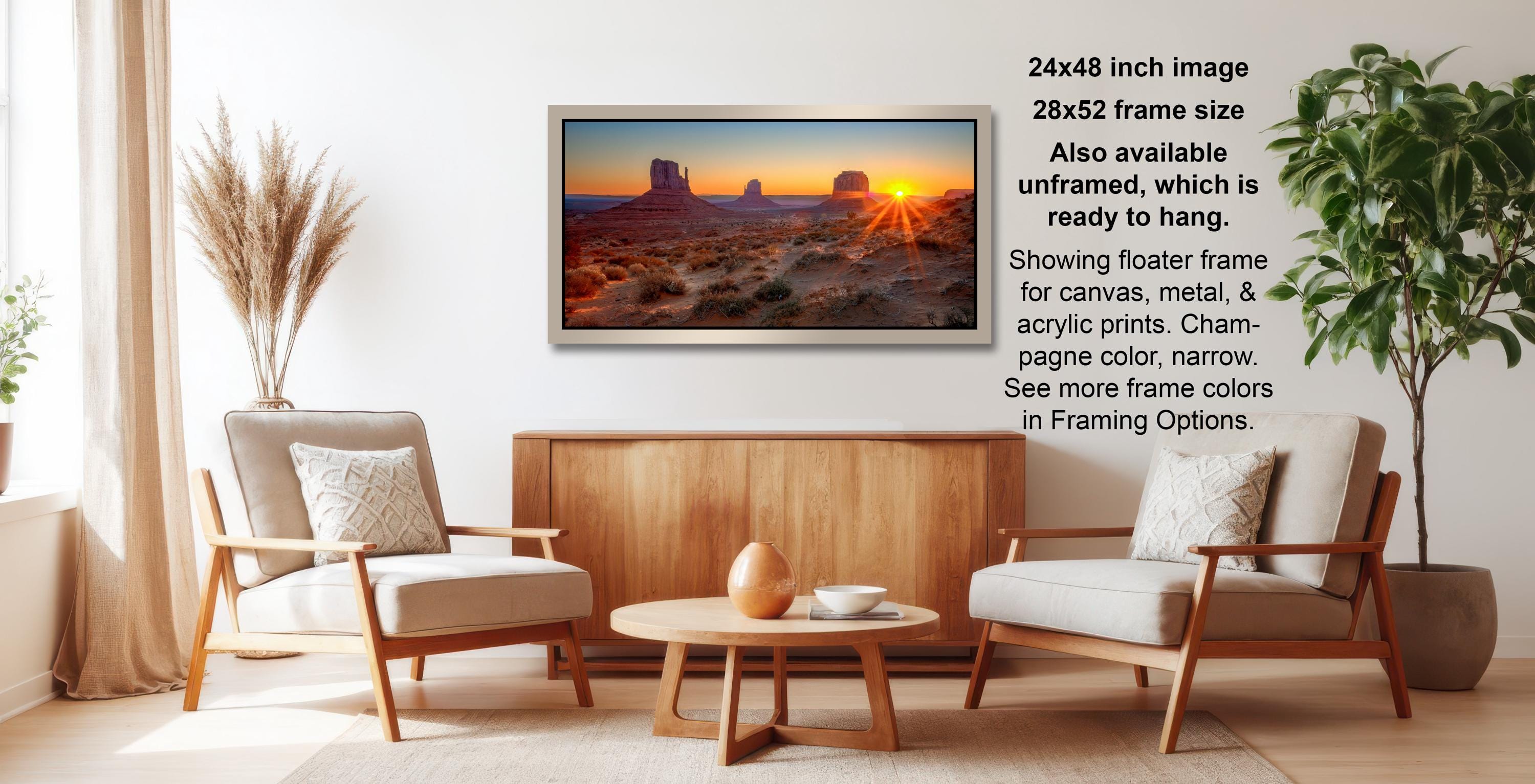 a living room with a large framed landscape photograph hanging on the wall. The photograph depicts a desert scene with a sunset, and the frame size is 24x48 inches. The frame is available in a 28x52-frame size, and it is ready to hang. The photograph is showing a floater frame for canvas, metal, and acrylic prints, with a charm-colored, narrow page color. The frame color options are listed, and the image also mentions that there are more frame colors in Framing Options.