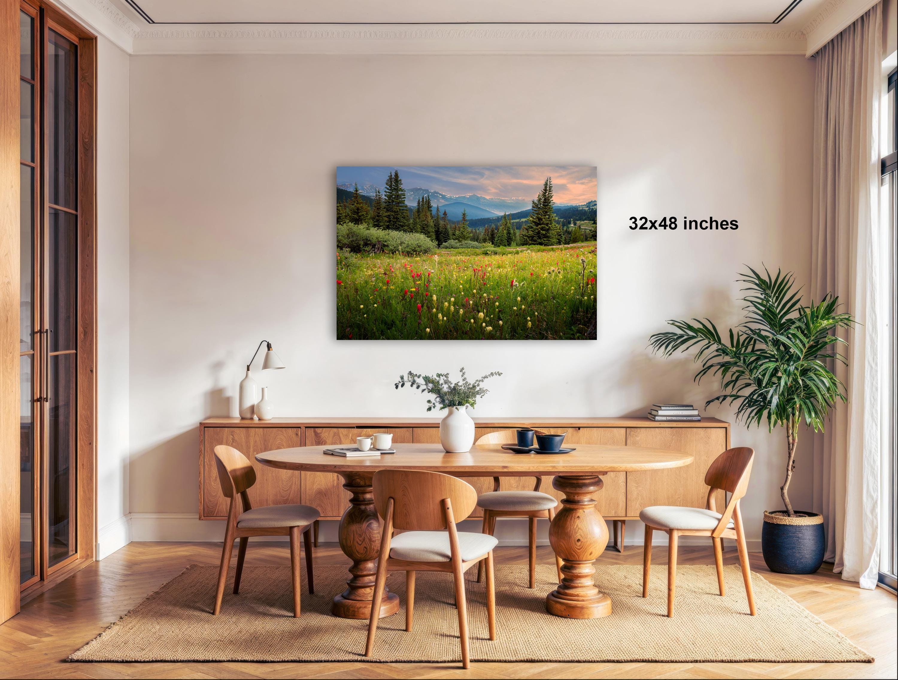 a serene dining room with a large wooden table surrounded by matching chairs. A painting of a field with wildflowers and mountains in the background is displayed on the wall above the table. The room is decorated with potted plants and other natural elements, creating a peaceful and inviting atmosphere.