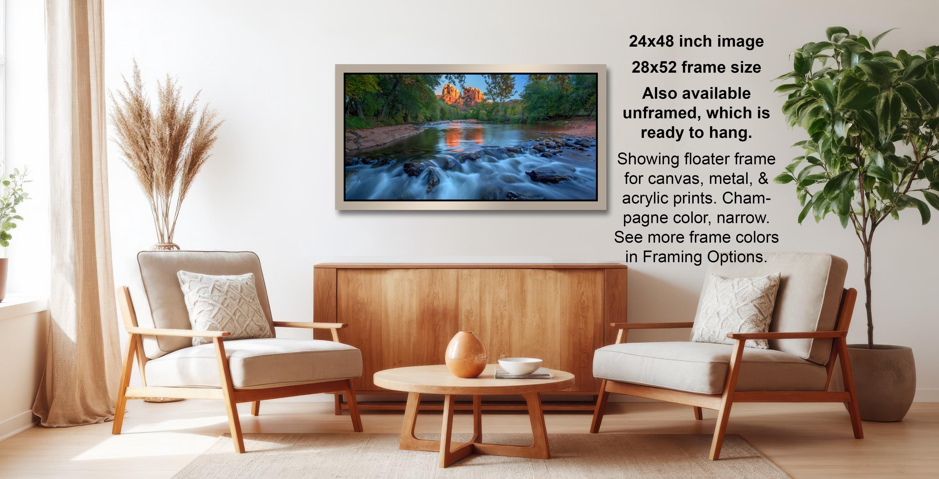 The image is a living room with a large framed landscape painting hanging on the wall. The painting depicts a serene river flowing through a forest, with a wooden fence and a bridge in the background. The room is furnished with two chairs and a coffee table, creating a cozy and inviting atmosphere.