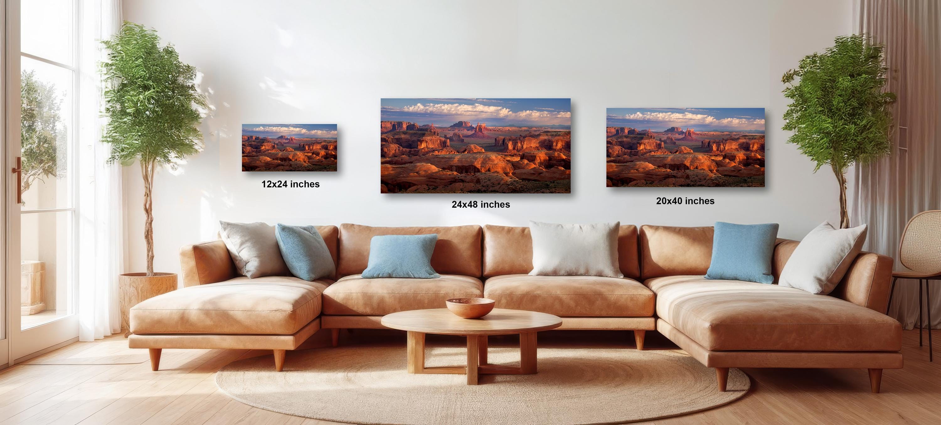 a large, brown sectional sofa with three framed landscape paintings hanging above it. The paintings depict desert scenes with mountains in the background. The sofa is positioned in front of a window, and there are two potted plants on either side of the sofa. The paintings are of different sizes, with the largest one being 24 inches wide and the smallest one being 12 inches wide.