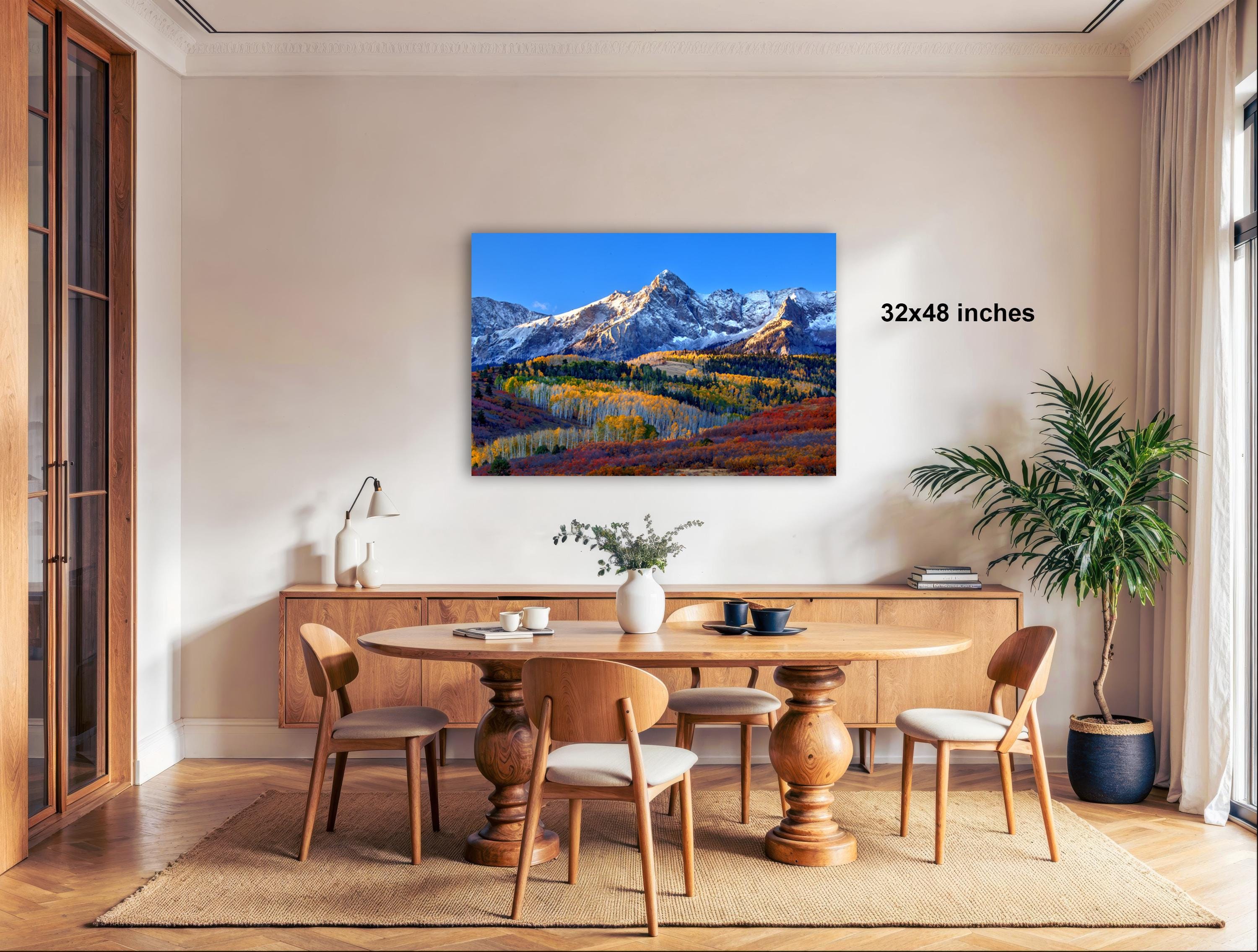 a large, framed landscape painting hanging on a wall in a modern dining room. The painting depicts a mountainous, autumnal scene with vibrant colors and a sense of tranquility. The dining table is surrounded by wooden chairs, and there are potted plants and other decorative elements in the room, creating a cozy and inviting atmosphere.