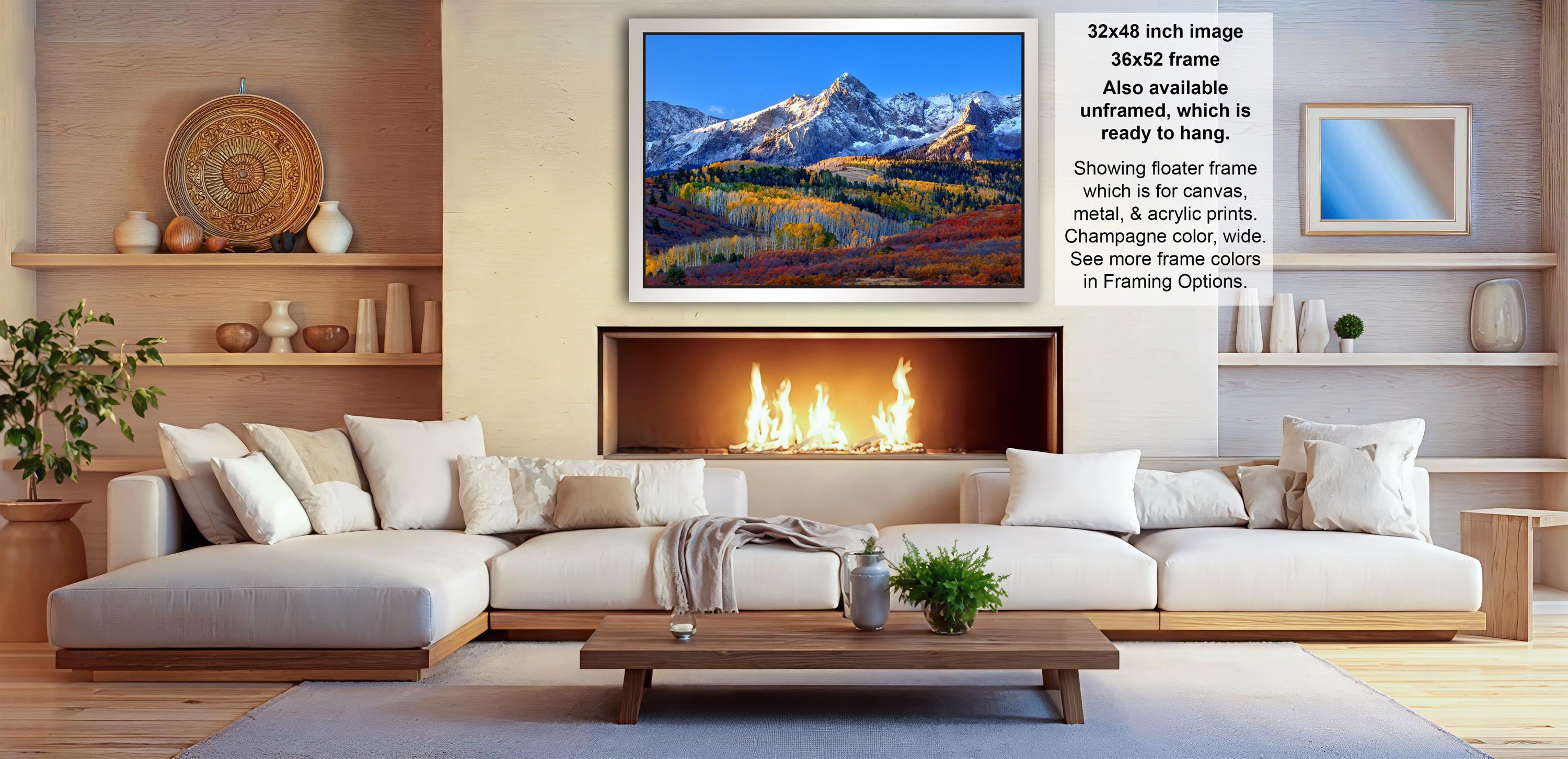 a cozy living room with a fireplace, white sofas, and a wooden coffee table. Above the fireplace, there is a framed picture of a mountain landscape. The room also features shelves with various decorative items, such as vases and a potted plant.