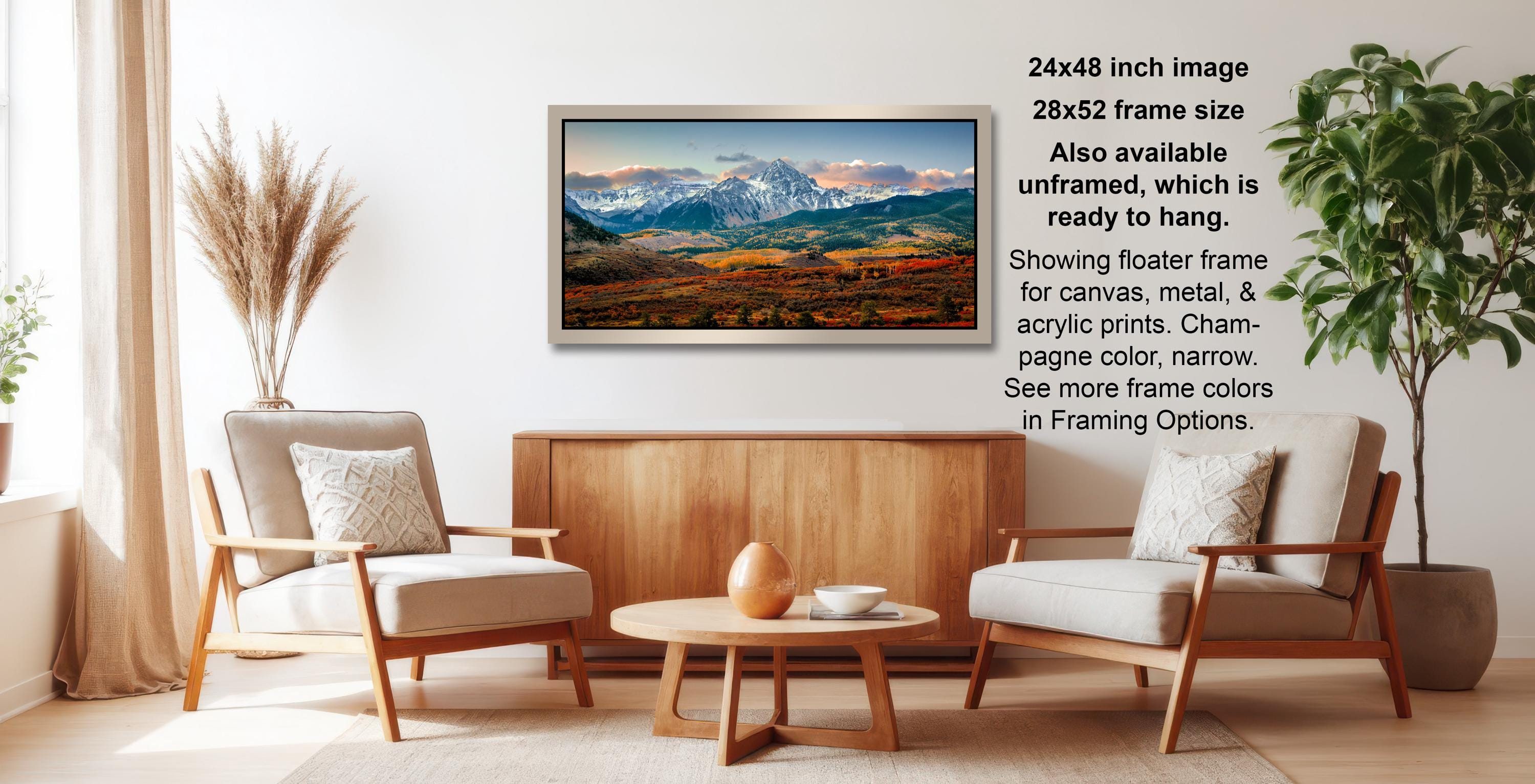 a living room with a large framed landscape painting hanging on the wall. The painting depicts a scenic mountain landscape with a clear blue sky and colorful foreground. The room is furnished with two armchairs and a coffee table, creating a cozy and inviting atmosphere.