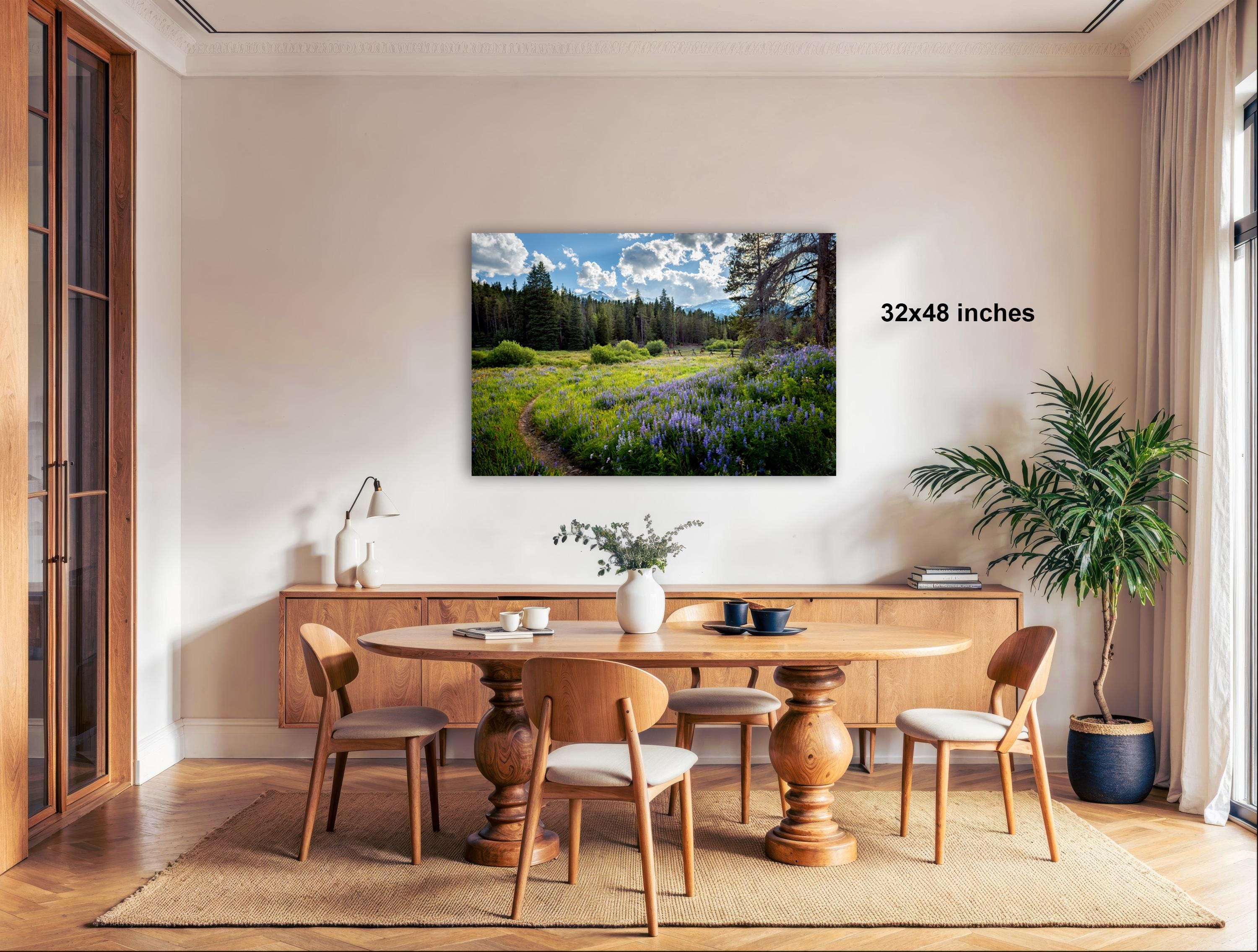 a serene dining room with a large wooden table surrounded by matching chairs. A painting of a field with purple flowers hangs on the wall above the table. The room is decorated with various plants and other items, creating a cozy and inviting atmosphere.