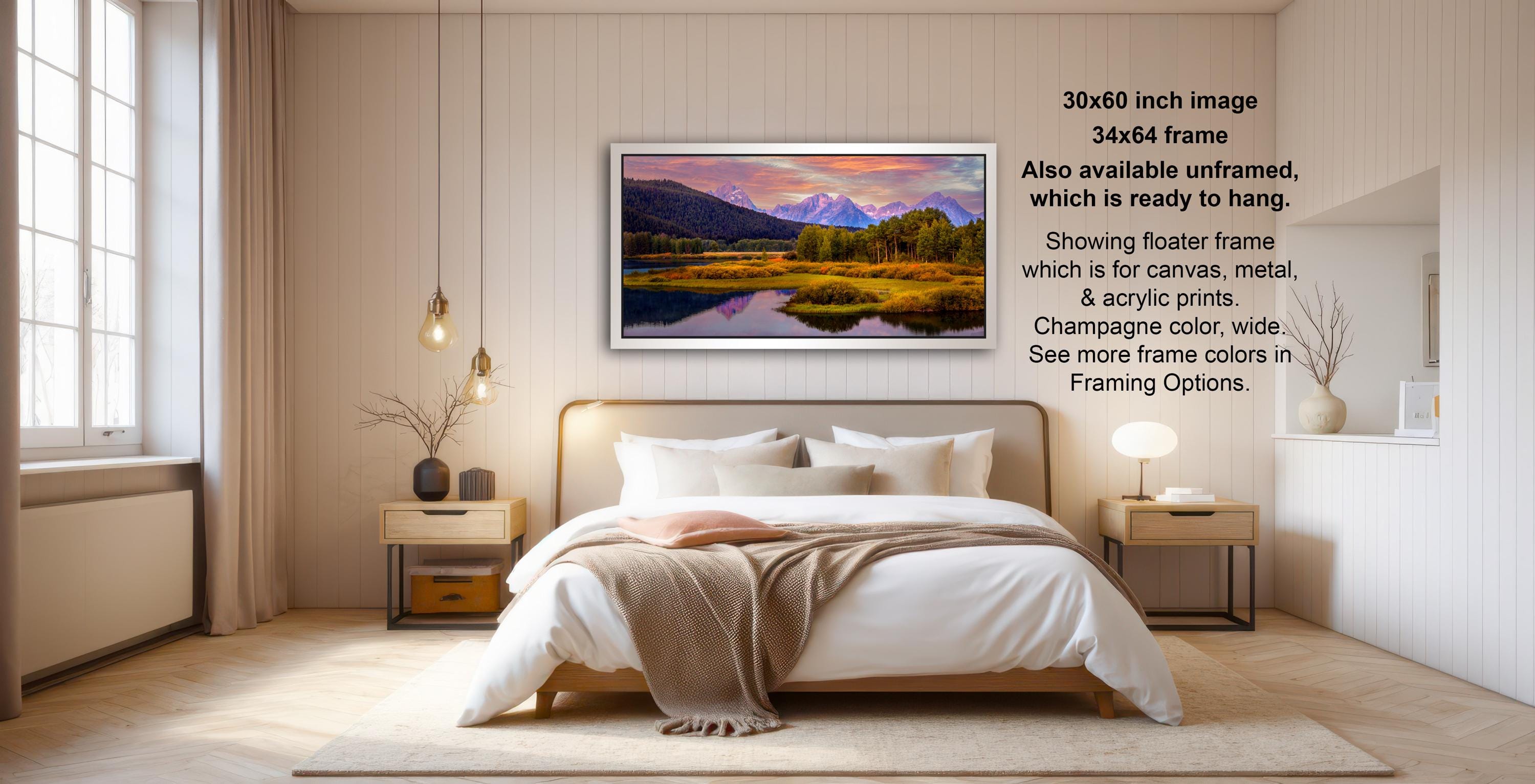 The image is a modern bedroom with a large bed, wooden furniture, and a framed landscape painting hanging on the wall. The painting depicts a serene mountain landscape with a lake and trees. The room has a minimalist and clean design, with a white color scheme and natural light coming in from a window.