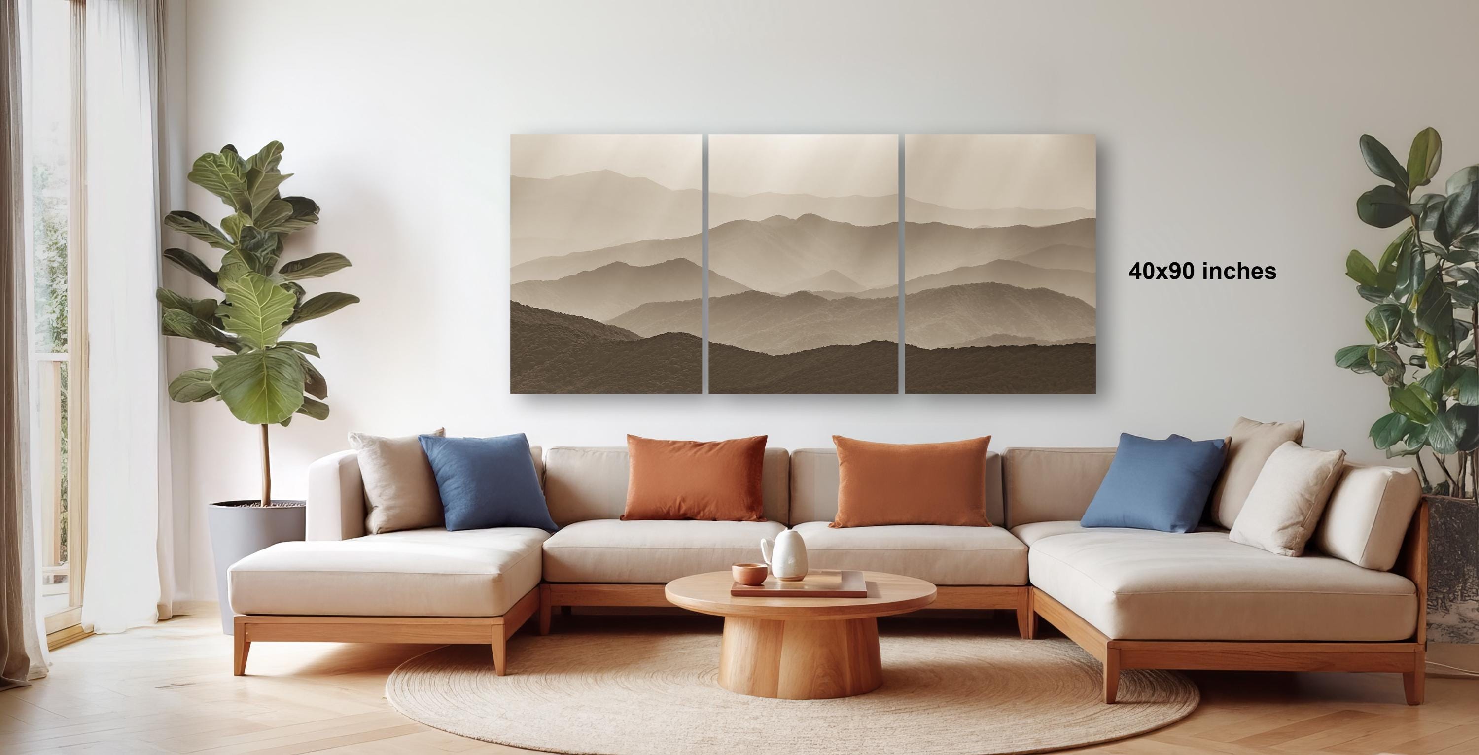 a modern living room with a large, three-panel landscape painting hanging on the wall above a beige sectional sofa. The sofa is adorned with colorful throw pillows, and there are two potted plants in the room. The overall decor has a minimalist and contemporary style.