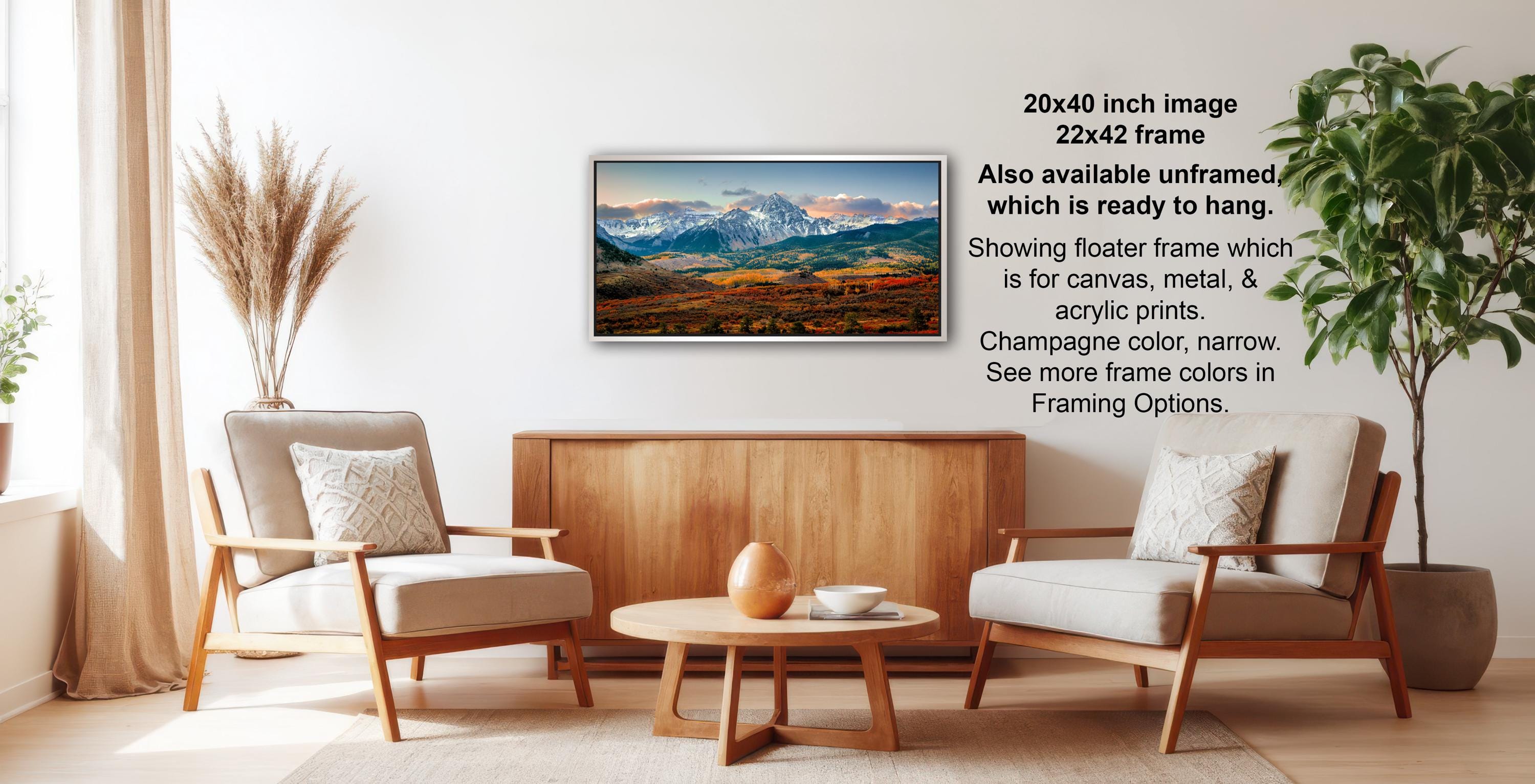 a living room with a wooden coffee table, two white armchairs, and a potted plant. A framed landscape painting is hanging on the wall, and there is text providing information about the frame options.
