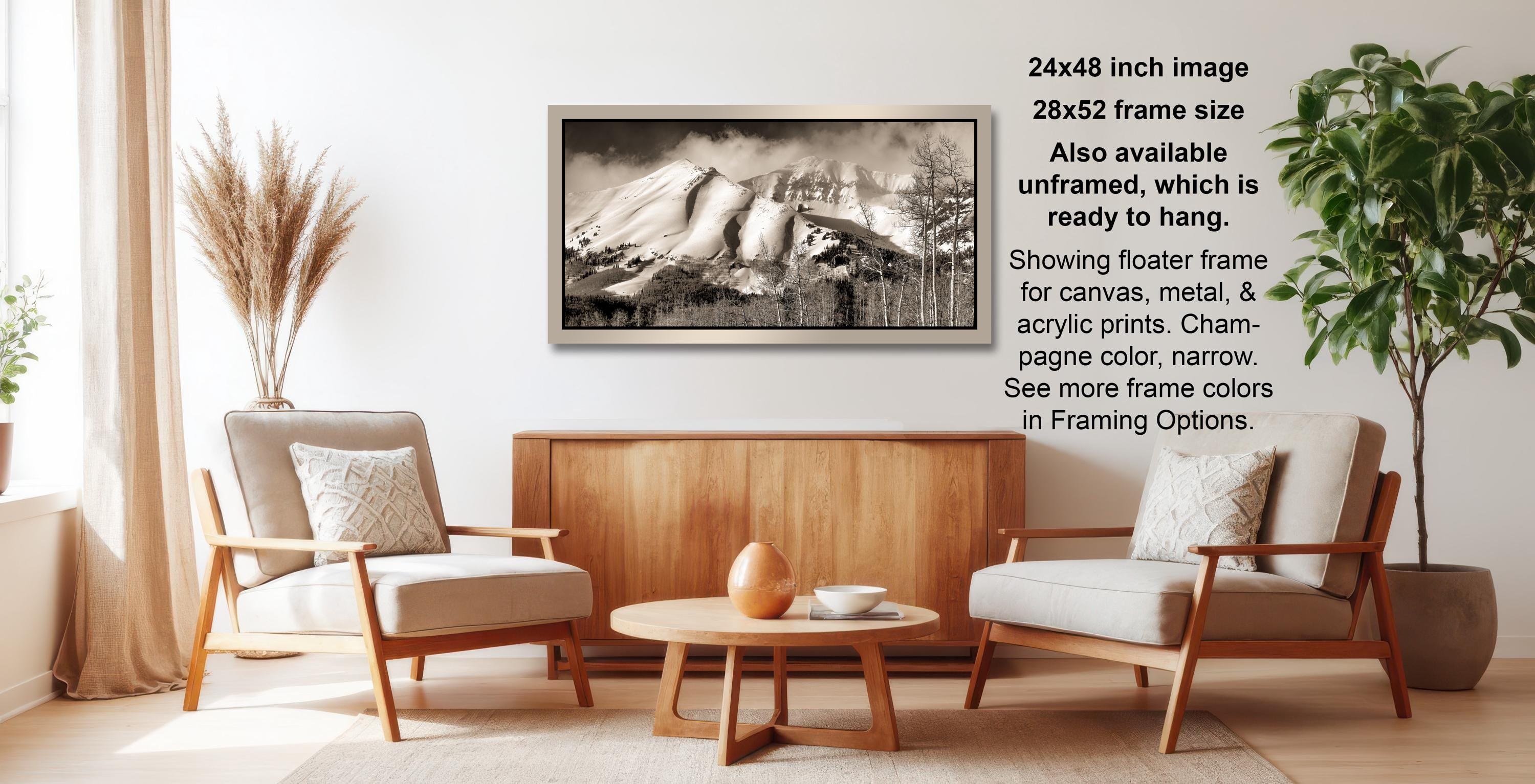 The image is a living room with a wooden coffee table, two white armchairs, and a framed black and white photograph of a mountain landscape hanging on the wall.