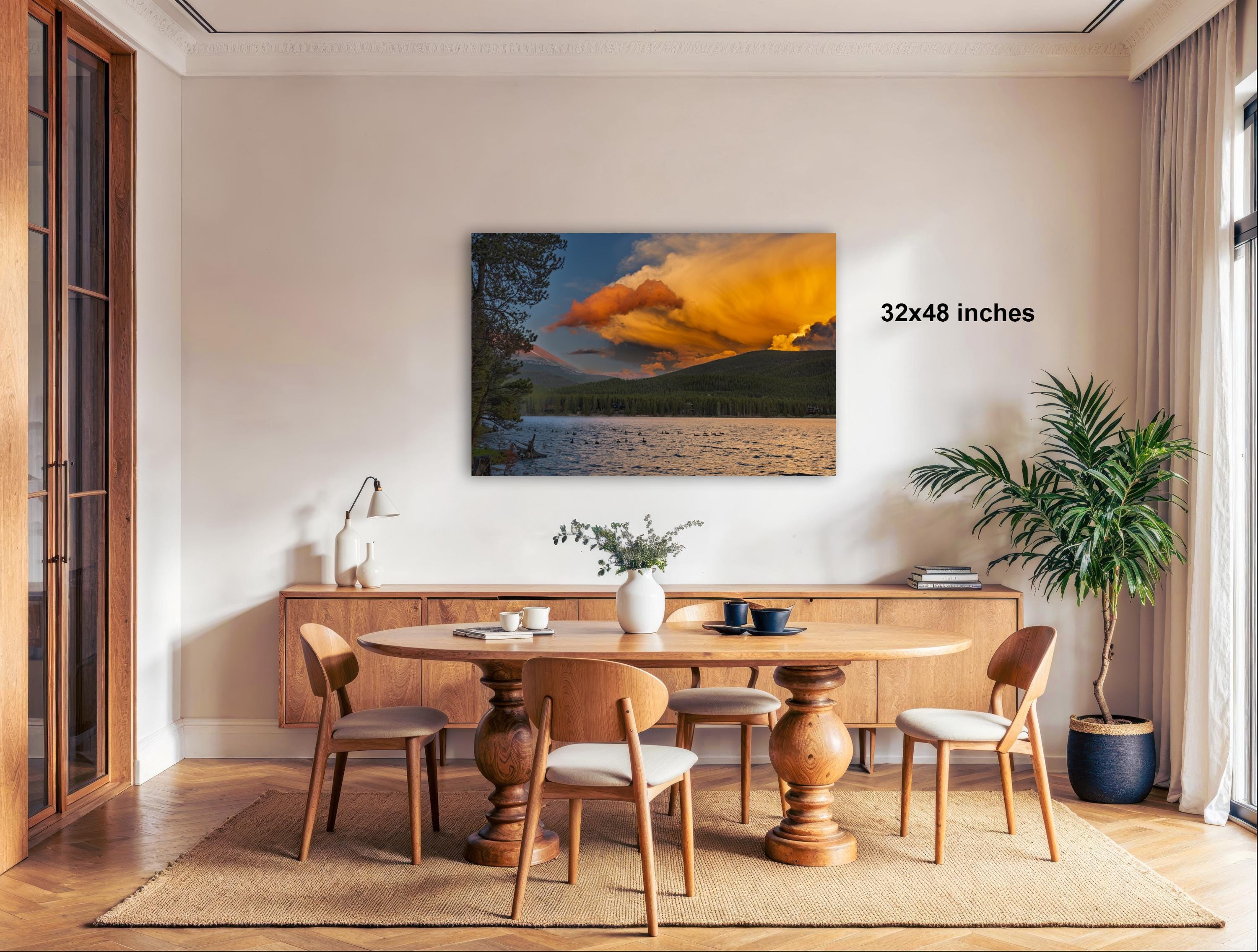 a modern dining room with a large wooden table surrounded by matching chairs. A large painting of a sunset landscape is displayed on the wall above the table. The room is decorated with various plants and other furnishings, creating a cozy and inviting atmosphere.