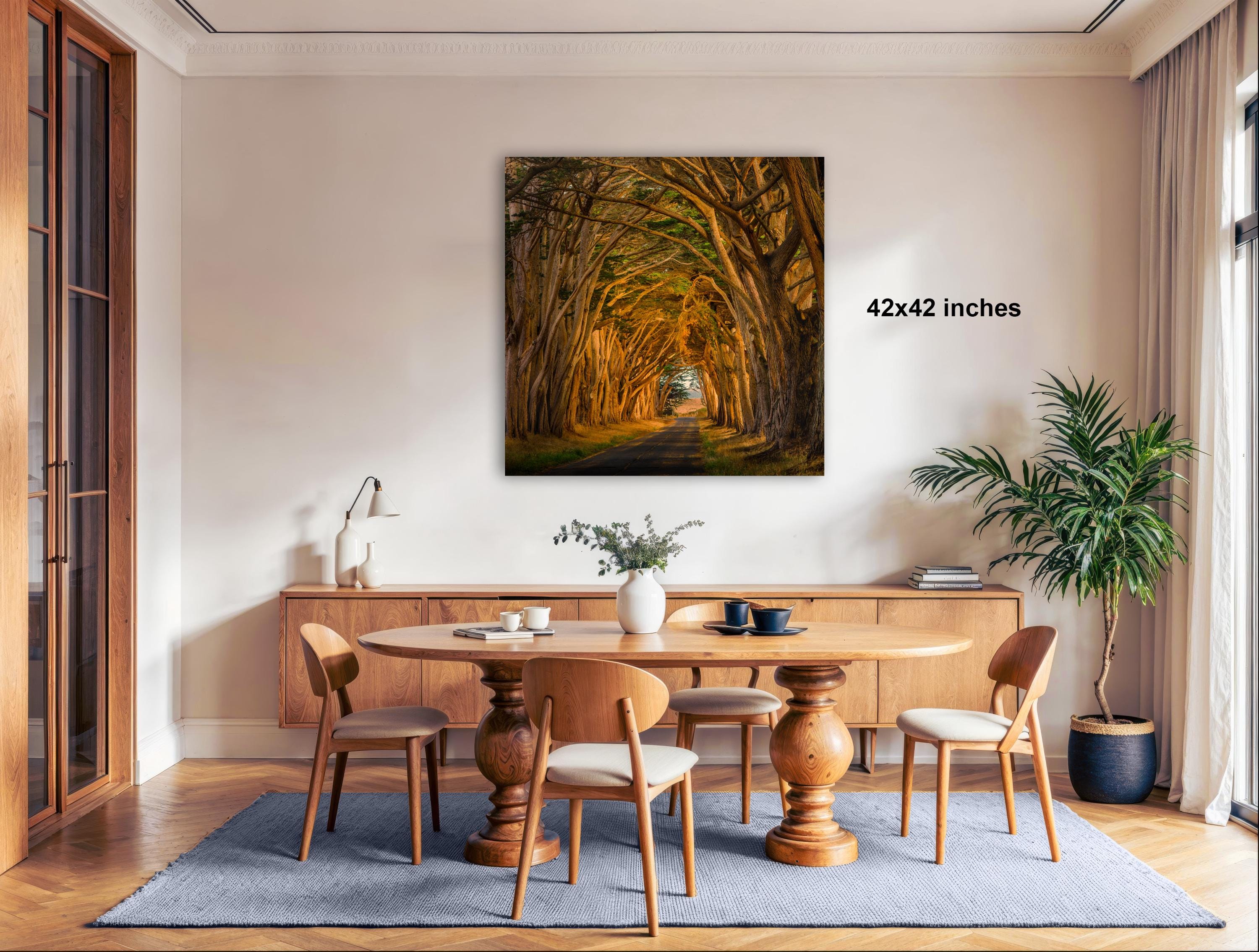 a serene dining room with a large wooden table surrounded by matching chairs. A large framed photograph of a tree-lined path is displayed on the wall above the table. The room is decorated with a potted plant, a vase, and a lamp, creating a cozy and inviting atmosphere.