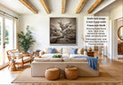 A living room with a large, framed black and white photograph of a tree on the wall. The room has a beige sofa, wooden chairs, and a large window overlooking a garden. The photograph appears to be of a desert landscape, with a sense of tranquility and natural beauty.