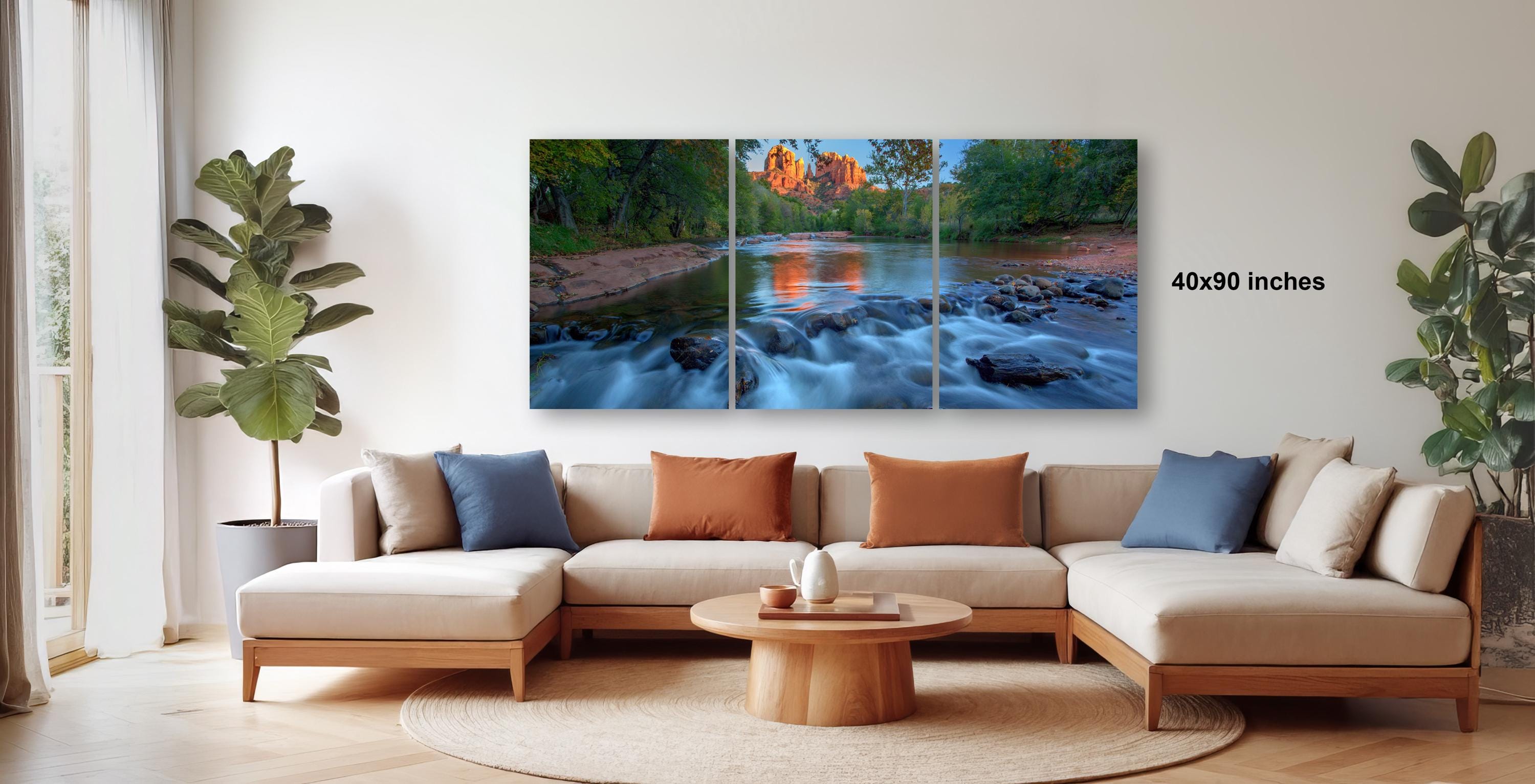 a modern living room with a large, colorful painting of a river and forest hanging on the wall. The painting is divided into three panels, showcasing the natural beauty of the landscape. The room is furnished with a beige sofa and a wooden coffee table, creating a cozy and inviting atmosphere. The room is decorated with potted plants and other natural elements, adding to the overall aesthetic.