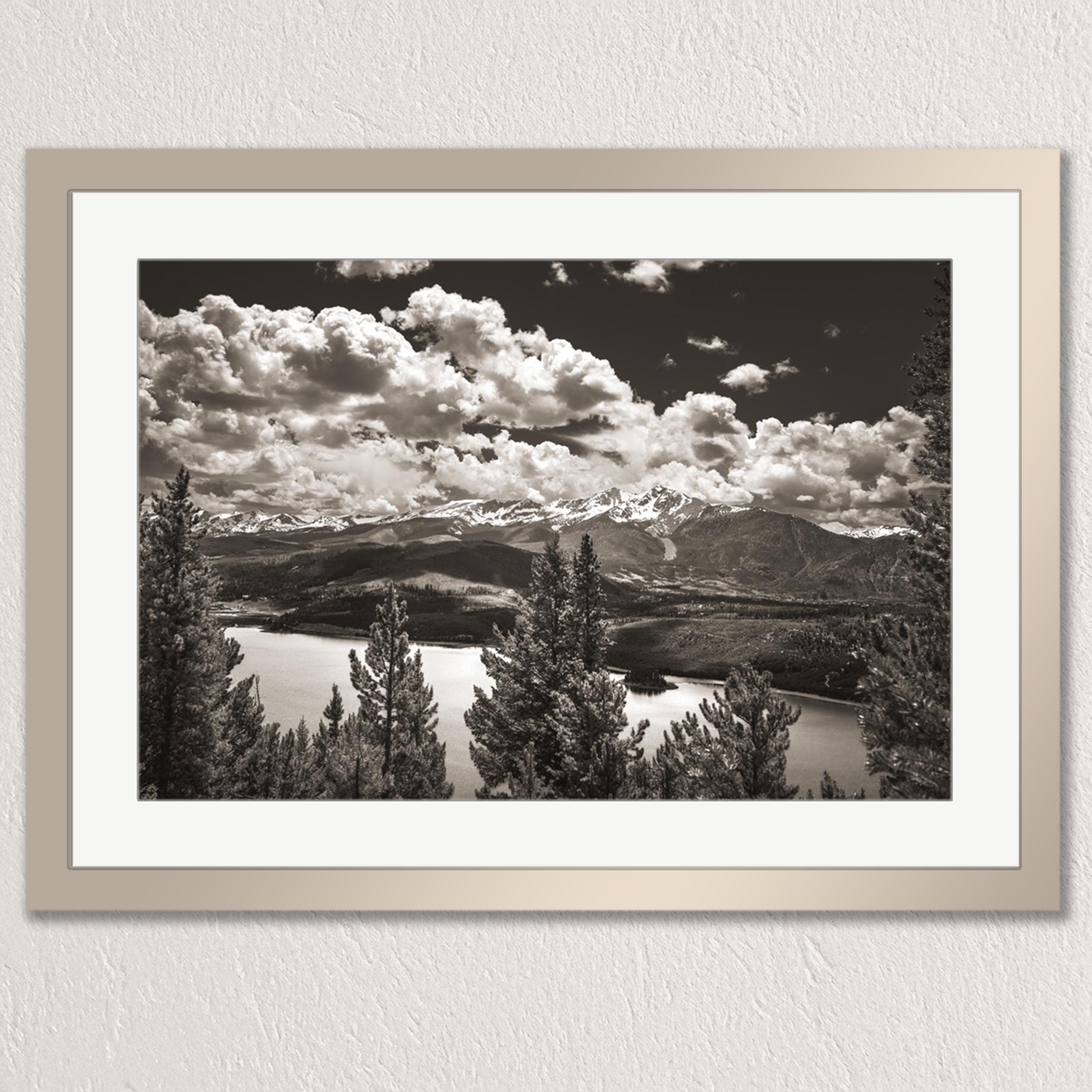 A black and white landscape photograph depicting a mountainous area with a lake or river in the foreground, surrounded by trees.