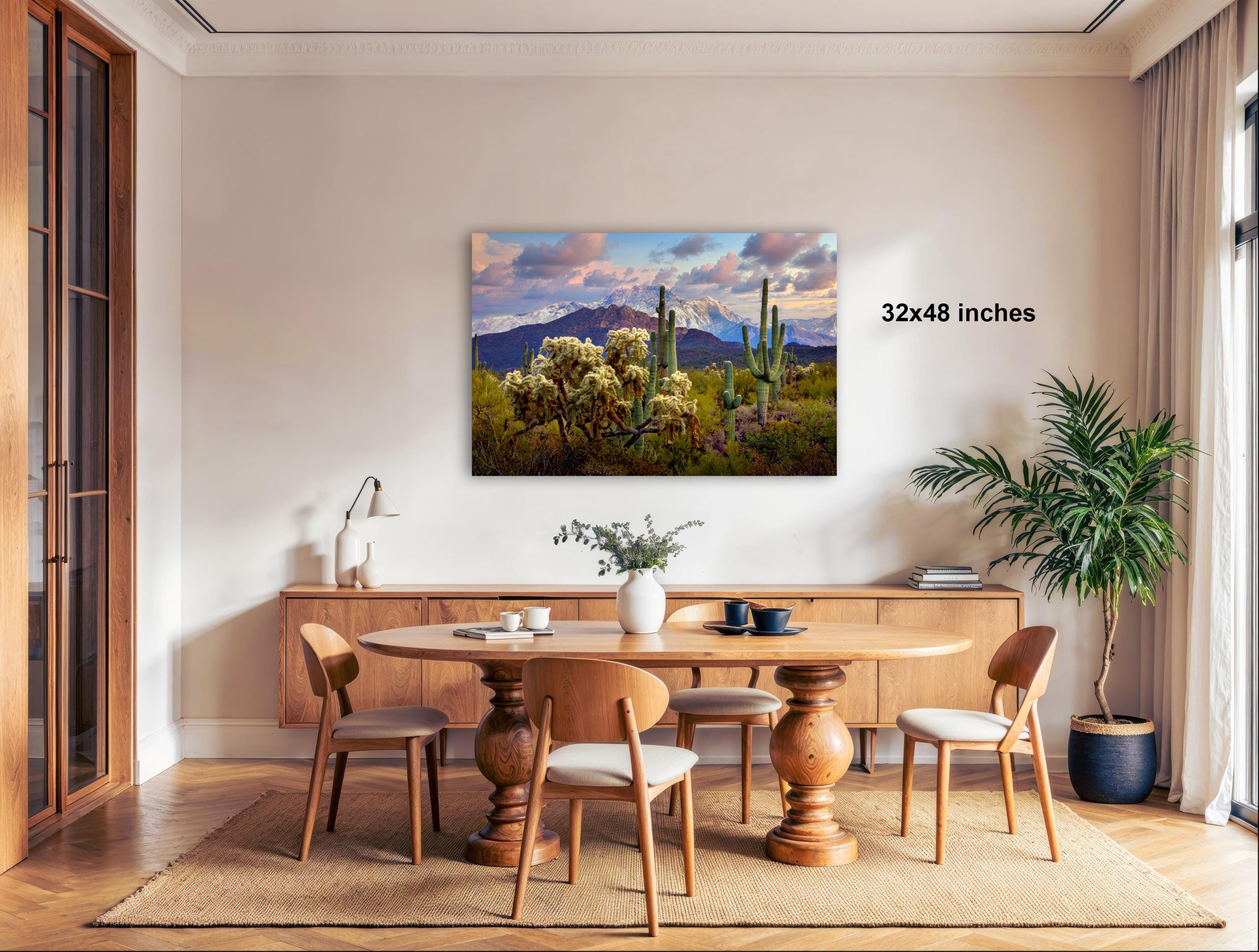 a serene desert landscape with a large cactus plant in the foreground, a wooden dining table surrounded by chairs, and a large framed print hanging on the wall.