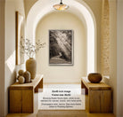The image is a black and white photograph of a hallway with a framed picture hanging on the wall, wooden tables, and decorative items such as vases and a bowl.