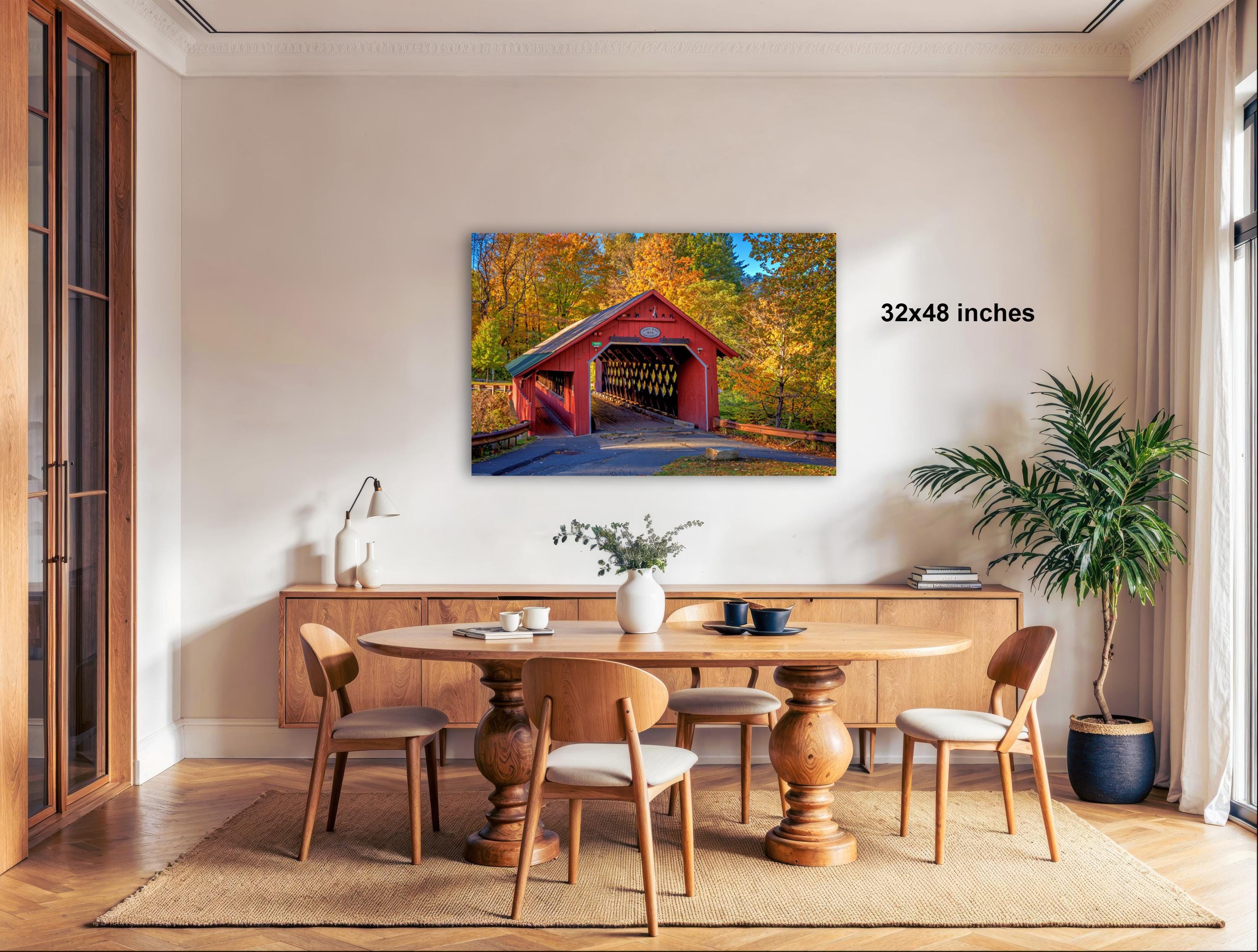 a cozy dining room with a wooden table and chairs, a potted plant, and a large framed photograph of a red covered bridge in the background.