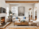 a cozy living room with a fireplace, a large framed picture of a forest, a white sofa, a coffee table, and various decorative elements such as potted plants and vases.