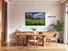 a serene dining room with a large wooden table surrounded by matching chairs. A framed landscape photograph of a field with trees and flowers is hanging on the wall above the table. The room is decorated with potted plants and other furnishings, creating a cozy and inviting atmosphere.