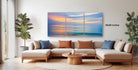 The image is a modern living room with a large, three-panel landscape painting hanging on the wall above a beige sectional sofa. The painting depicts a serene sunset over the ocean, with warm hues of orange, pink, and blue. The room is decorated with potted plants and a wooden coffee table, creating a cozy and inviting atmosphere.
