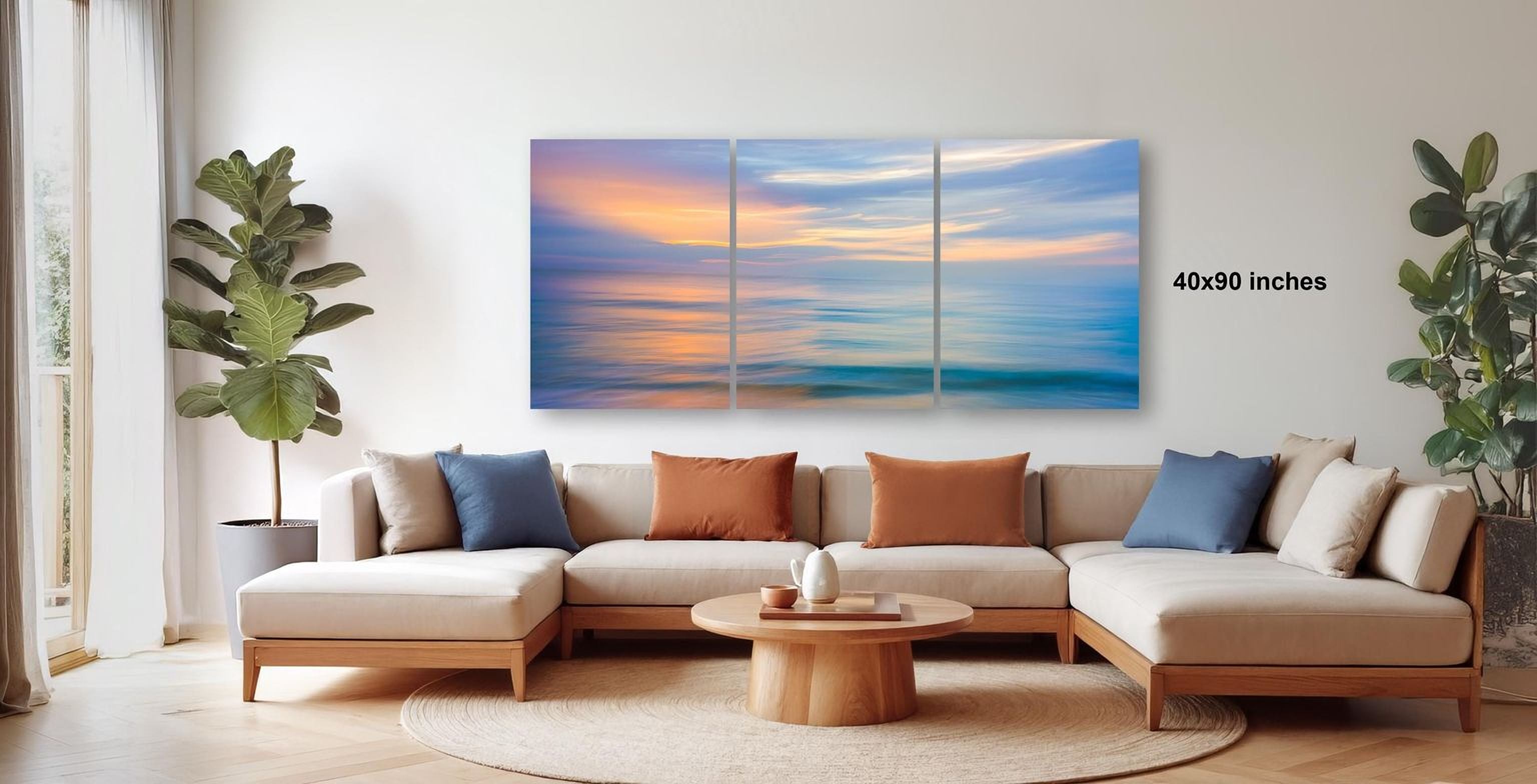 The image is a modern living room with a large, three-panel landscape painting hanging on the wall above a beige sectional sofa. The painting depicts a serene sunset over the ocean, with warm hues of orange, pink, and blue. The room is decorated with potted plants and a wooden coffee table, creating a cozy and inviting atmosphere.