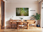 a serene dining room with a large wooden table surrounded by matching chairs. A painting of a forest path is displayed on the wall above the table, adding a natural element to the space. The room is well-lit, with natural light coming in from a window on the right side. The overall atmosphere is calm and inviting, with a touch of greenery from the potted plant and the vase of flowers on the table.
