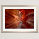 The image is a framed photograph of a striking natural rock formation, with a vibrant red and orange color palette that creates a sense of depth and movement. The photograph is framed in a white border, which contrasts with the warm tones of the rock formation. The image does not contain any discernible text or specific actions, and it is a single, cohesive image focused on the captivating rock formation.