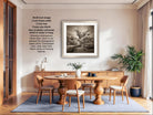 The image is a modern dining room with a wooden table and chairs, a potted plant, and a framed black and white photograph of a tree on the wall.