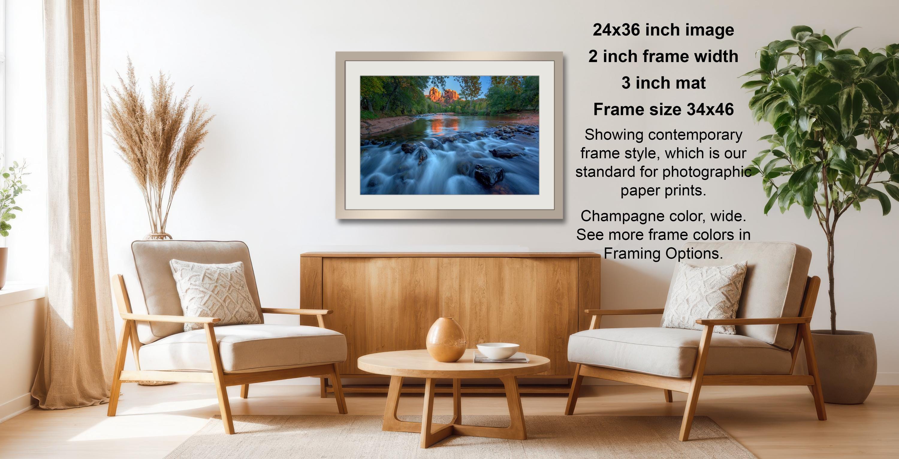 a living room with a wooden coffee table, two white armchairs, and a framed photograph of a river with a waterfall. The photograph is framed in a 2x3 grid format and has a size of 34x46 inches. The frame color is champagne, and the mat is 3 inches wide. The photograph is showing a contemporary frame style, which is the standard for photographic paper prints. The image also mentions that the frame color is wide and that there are more frame colors available in Framing Options.