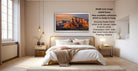 The image is a bedroom with a large bed, a nightstand, and a painting of a mountain landscape hanging on the wall. The painting is framed in a wooden frame and is ready to hang. The room has a minimalist and modern design, with a white color scheme and a neutral color palette.