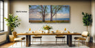 a dining room with a large wooden table set for four, surrounded by four chairs. Above the table, there is a large painting depicting a beach scene with trees and the ocean. The painting is framed and hangs on the wall, creating a serene and inviting atmosphere in the room.