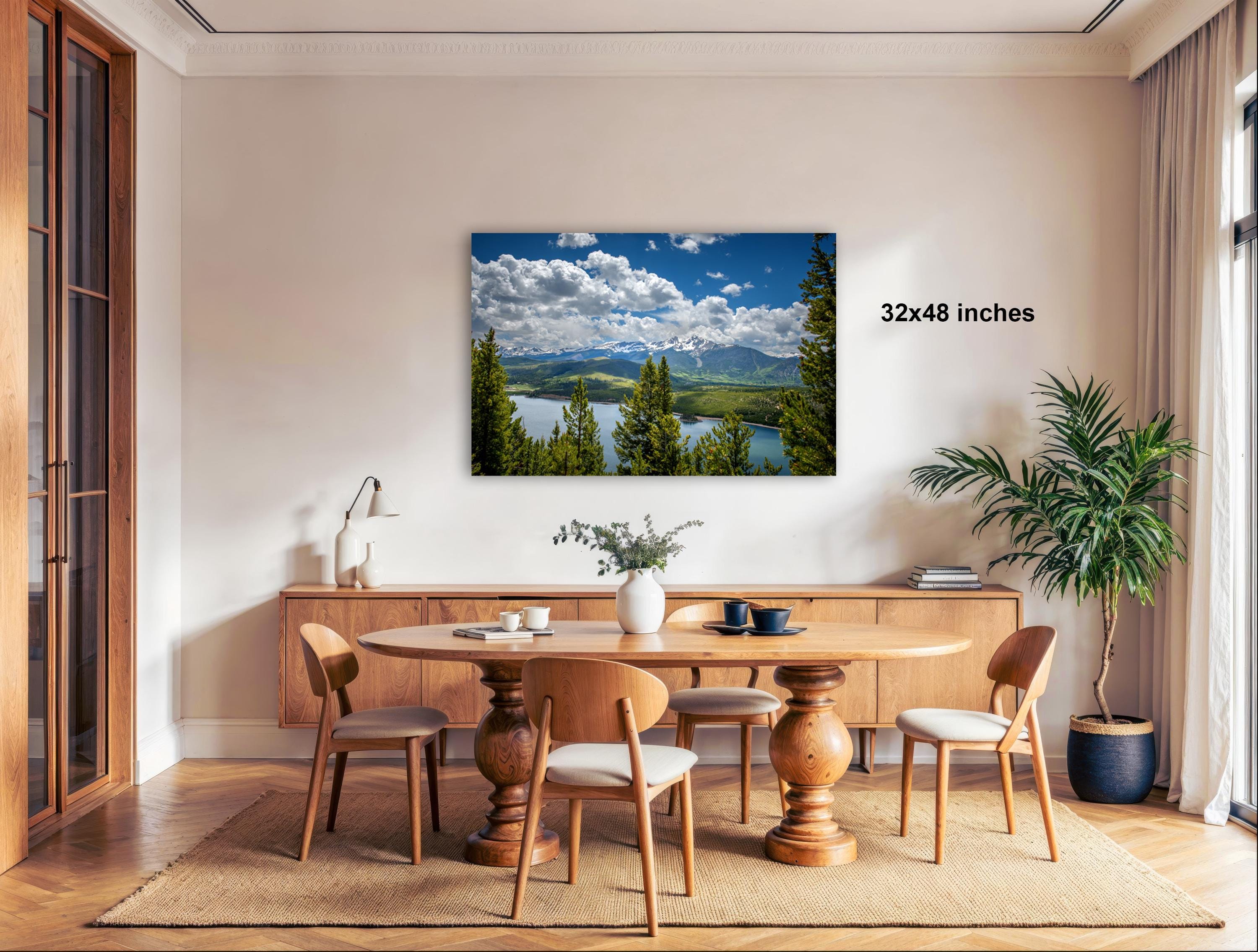 a dining room with a large wooden table surrounded by chairs, a potted plant, and a framed landscape photograph hanging on the wall.