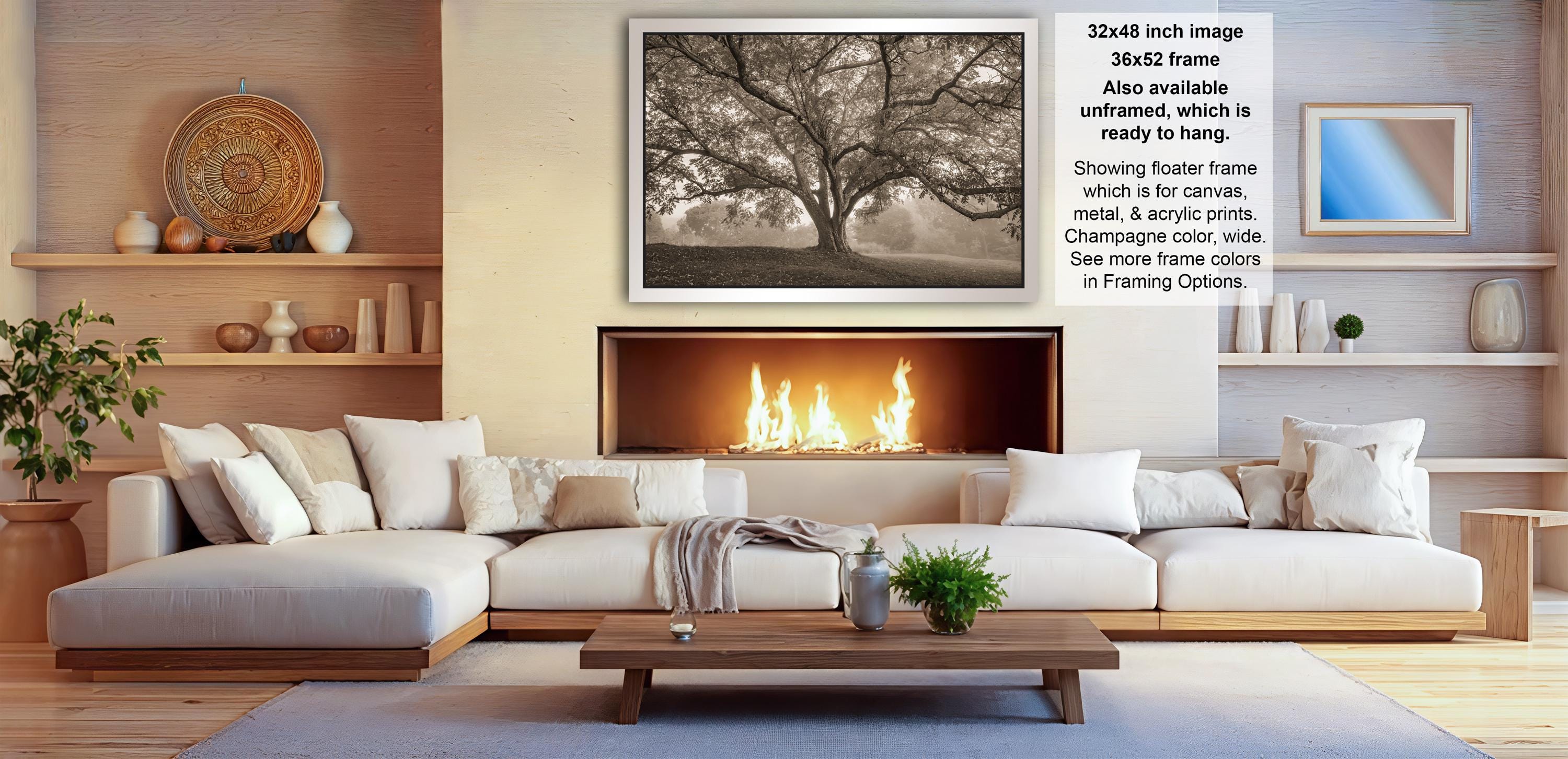 a cozy living room with a large white sectional sofa, a fireplace, and a wooden coffee table. Above the sofa, there is a framed photograph of a tree with a fluttering flag. The room also has shelves displaying various decorative items, including vases and a potted plant.