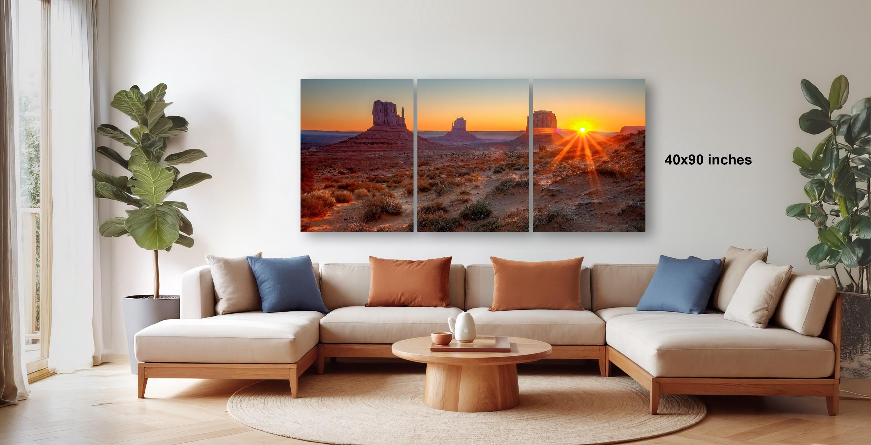 The image is a modern living room with a large, three-panel landscape painting depicting a desert scene at sunset. The painting is framed and hangs on the wall above a beige sectional sofa and a wooden coffee table.