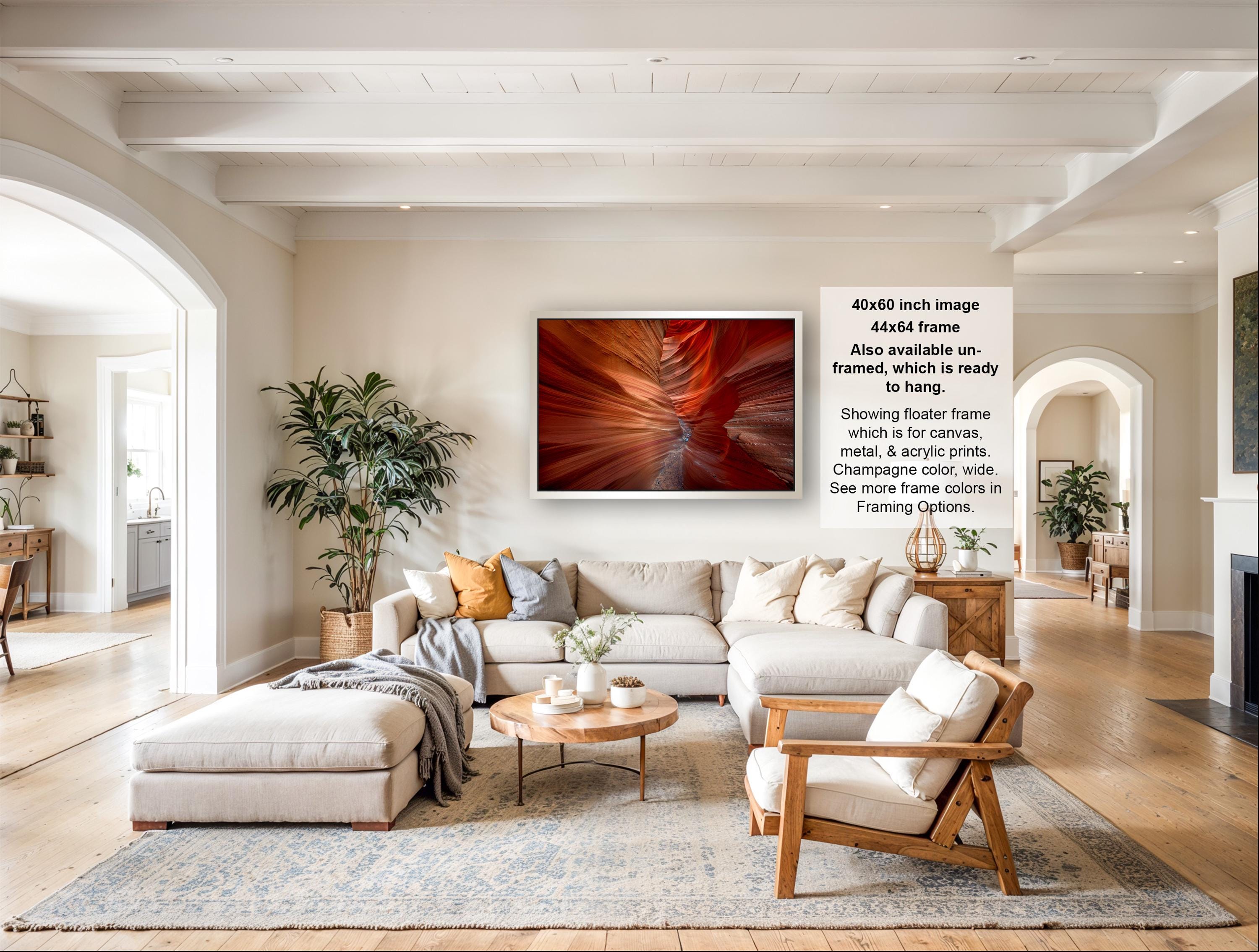 The image is a large, high-quality photograph of a living room with a large, framed artwork depicting a canyon or desert landscape. The room has a neutral color scheme with white walls, a gray rug, and a wooden coffee table. The furniture includes a sofa, armchairs, and a wooden side table. The room also features several potted plants and decorative items, creating a cozy and inviting atmosphere.