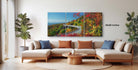 The image is a modern living room with a large, colorful painting of a scenic mountain landscape hanging on the wall. The painting features a winding road surrounded by trees and foliage in various shades of green, red, and orange. The room is furnished with a beige sofa and a wooden coffee table, creating a cozy and inviting atmosphere.