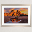 a stunning desert landscape with a large rock formation in the background, its orange and red hues contrasting beautifully with the vibrant colors of the sky. The rock formation is reflected in a small pool of water, creating a mirror image that adds depth and dimension to the scene. The image is framed and displayed on a wall, showcasing the captivating beauty of the desert landscape.
