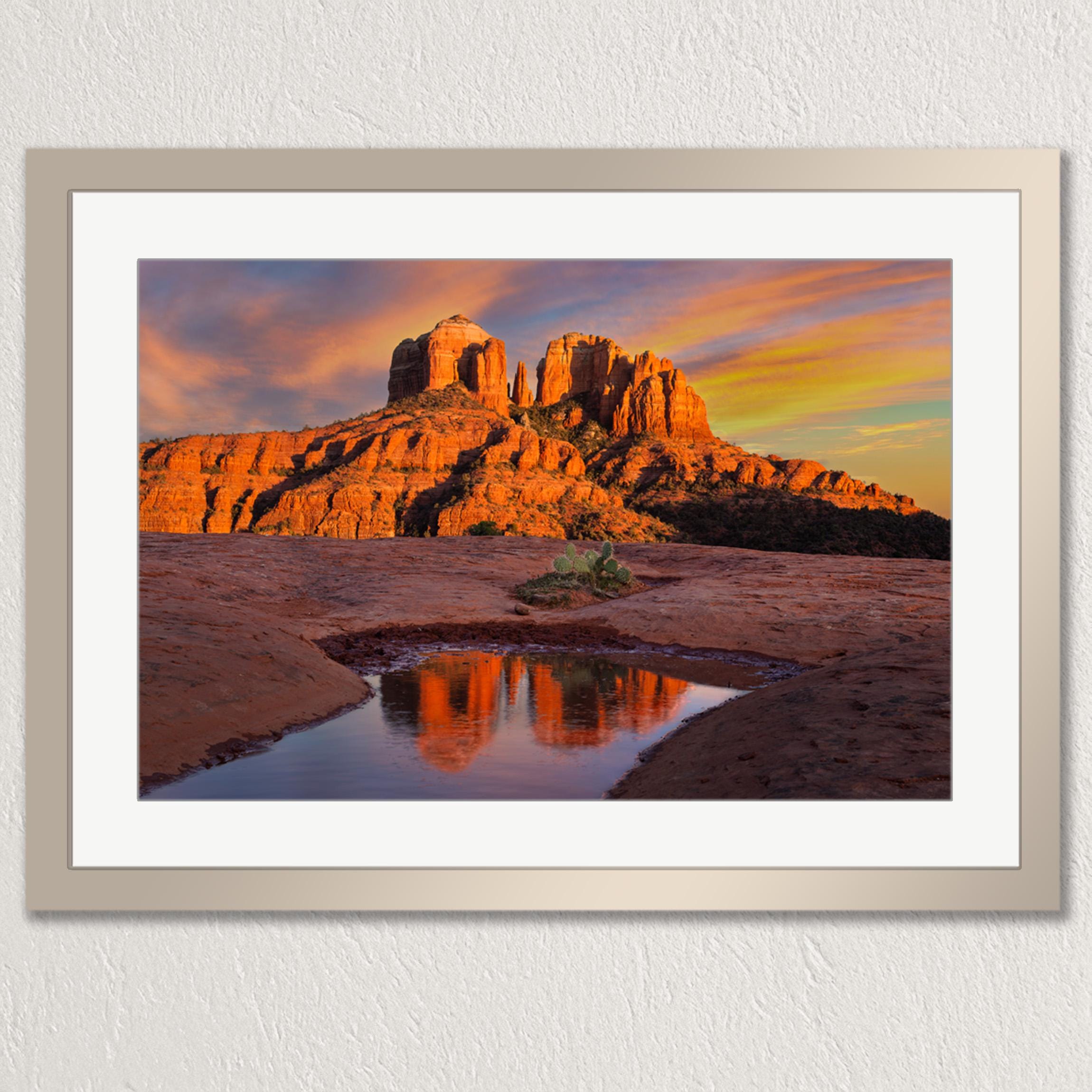 a stunning desert landscape with a large rock formation in the background, its orange and red hues contrasting beautifully with the vibrant colors of the sky. The rock formation is reflected in a small pool of water, creating a mirror image that adds depth and dimension to the scene. The image is framed and displayed on a wall, showcasing the captivating beauty of the desert landscape.