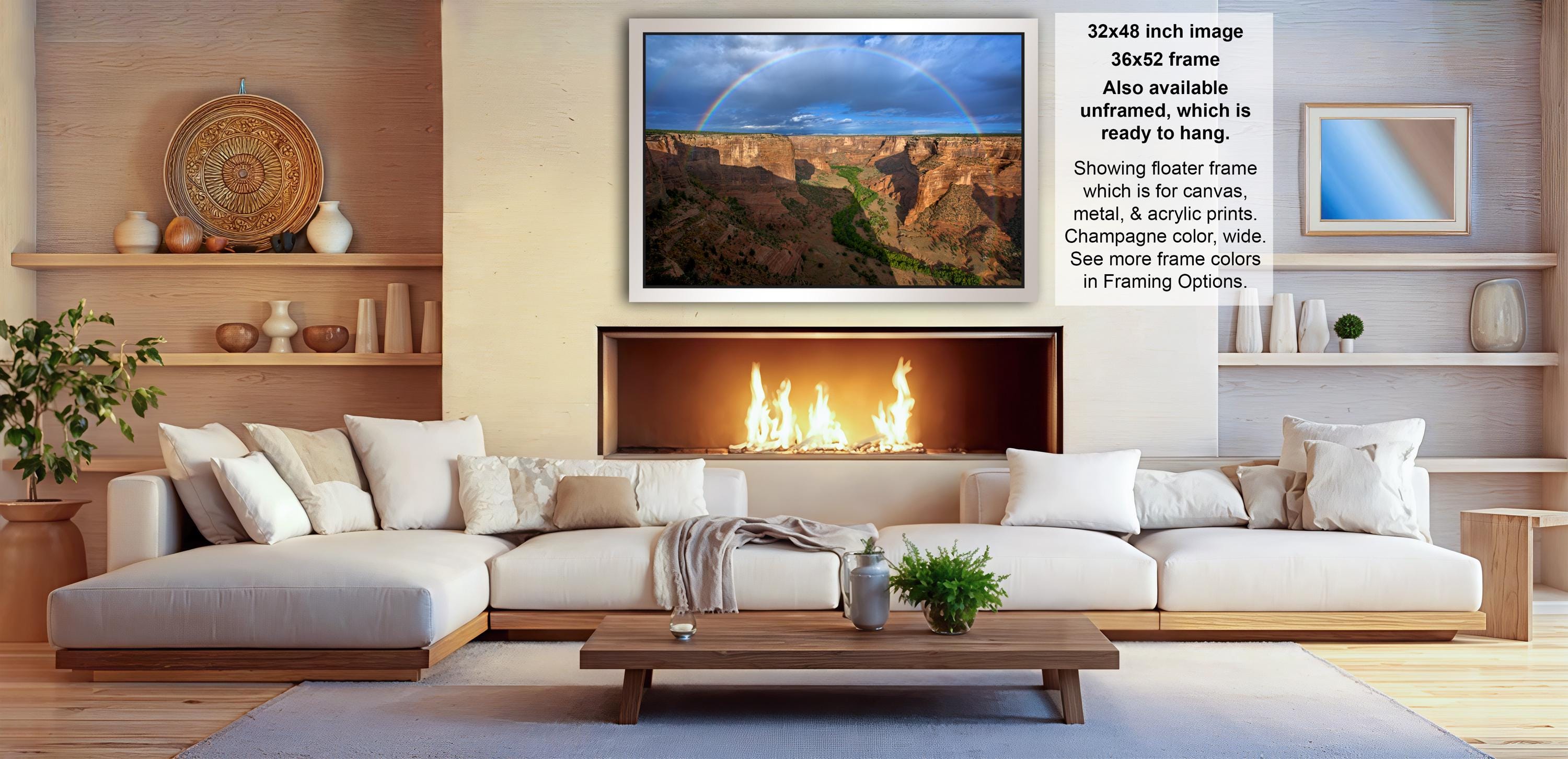 a cozy living room with a fireplace, white sofas, and a wooden coffee table. Above the fireplace, there is a framed image of a canyon with a rainbow in the sky. The room also has shelves displaying various decorative items, such as vases and a potted plant.