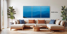 a modern living room with a large, three-panel landscape painting hanging on the wall above a beige sectional sofa. The painting depicts a serene mountain landscape with a bright orange sun in the sky. The room is decorated with potted plants and a wooden coffee table, creating a cozy and inviting atmosphere.