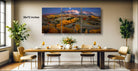 a large, multi-panel landscape painting hanging on a wall above a wooden dining table set with wine glasses, plates, and a vase of flowers.
